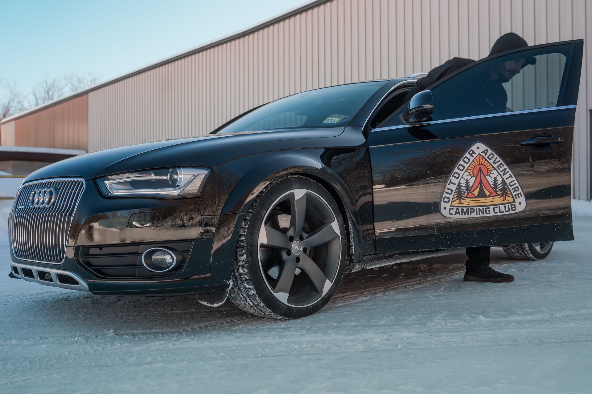 Black Audi with a person exiting it on a snowy surface; a logo is visible on the door.
