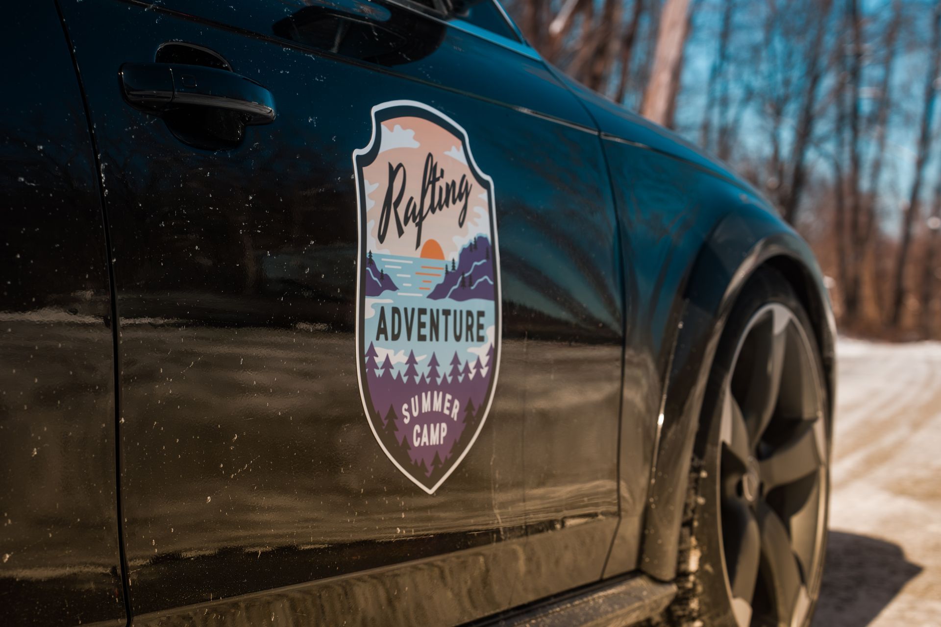 Black car door with a “Rolling Adventure Camp” badge on a sunny dirt road, trees in the background