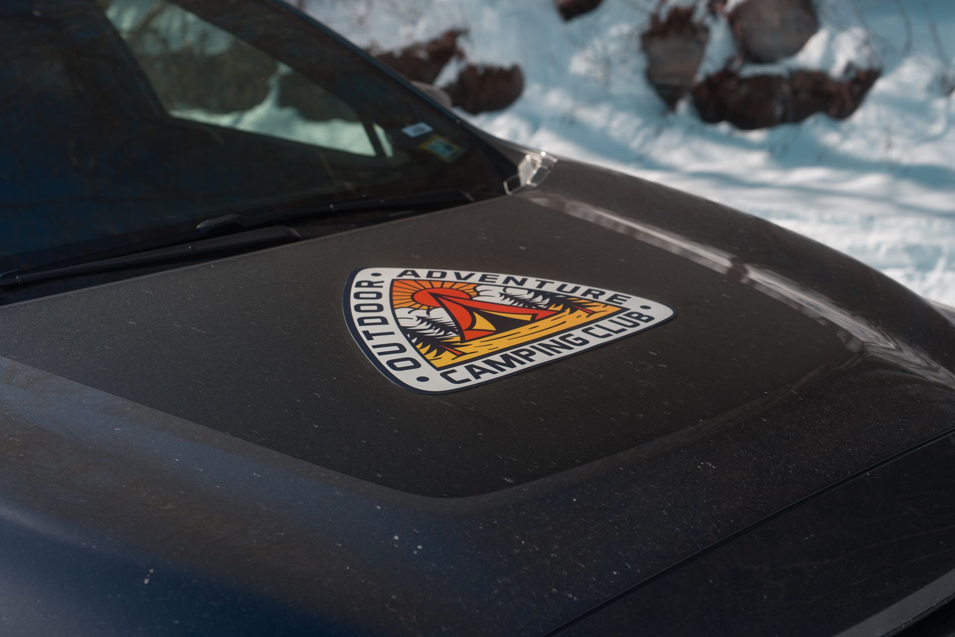 Close-up of a black car hood with a colorful sticker, parked in snowy surroundings