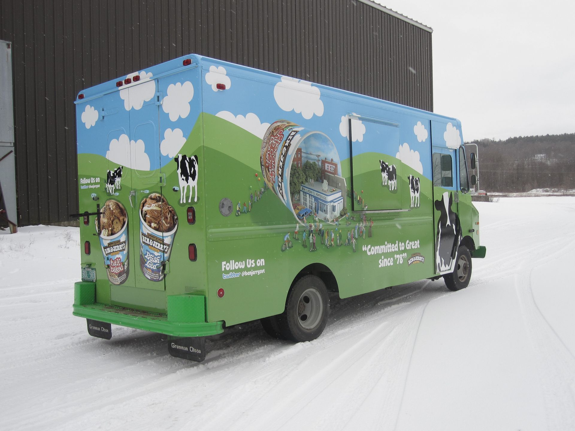 Green ice cream truck with cow graphics driving on a snowy road beside a building