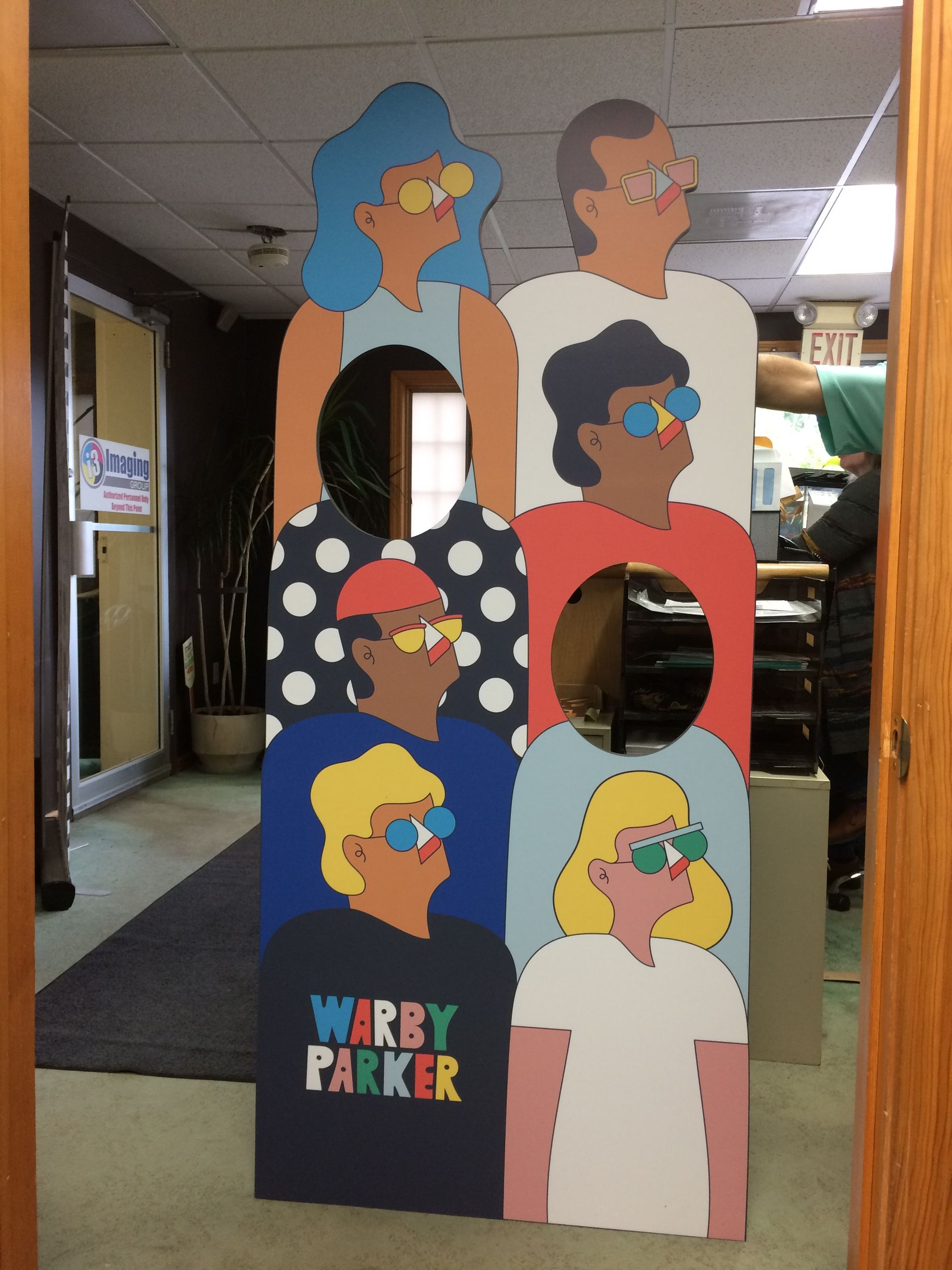 Cardboard standee with colorful cartoon faces and two face cutouts in a hallway, labeled “Wavy Parker”