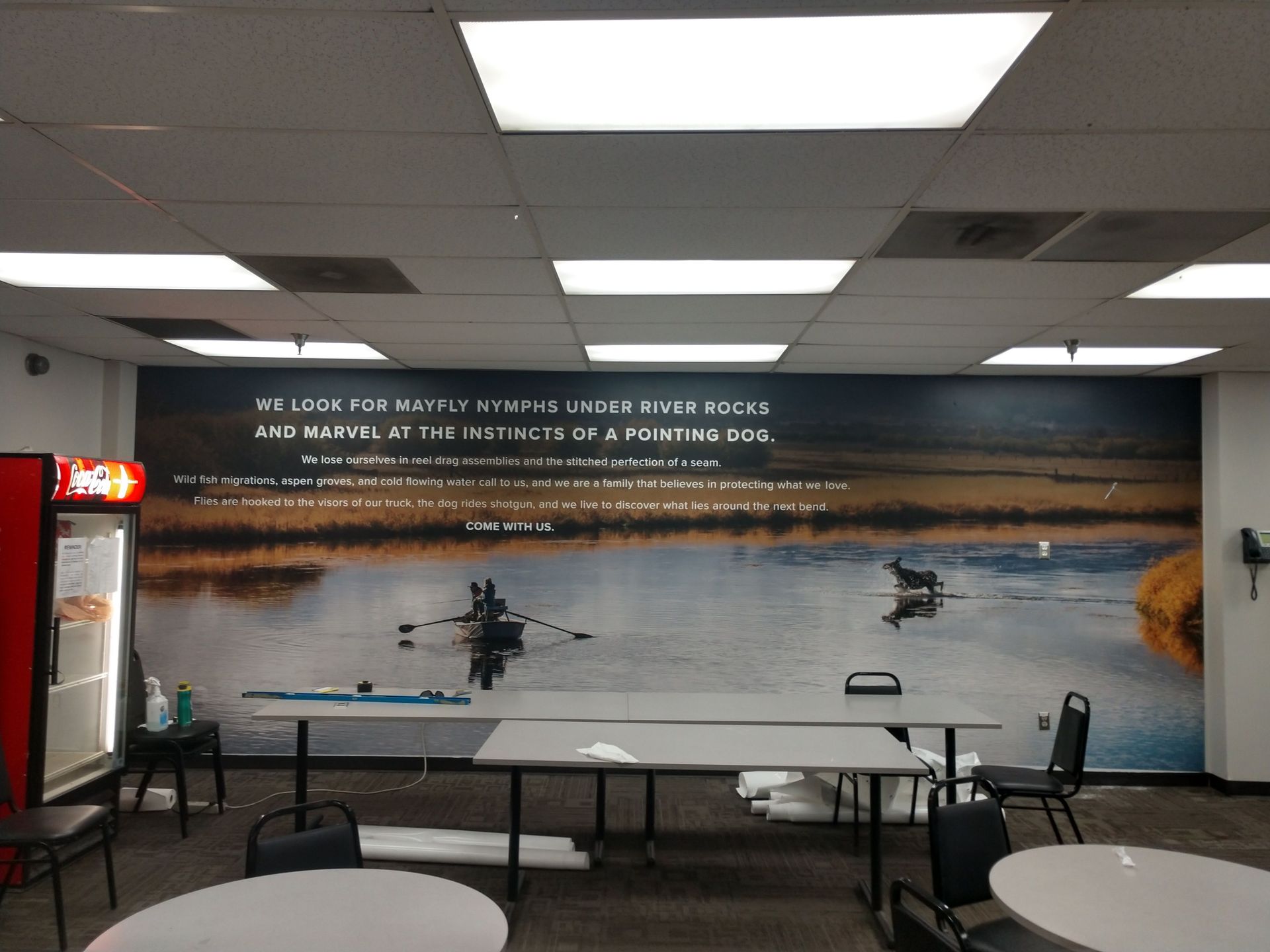 Indoor lounge with tables and chairs facing a large wall mural of a lake at sunset