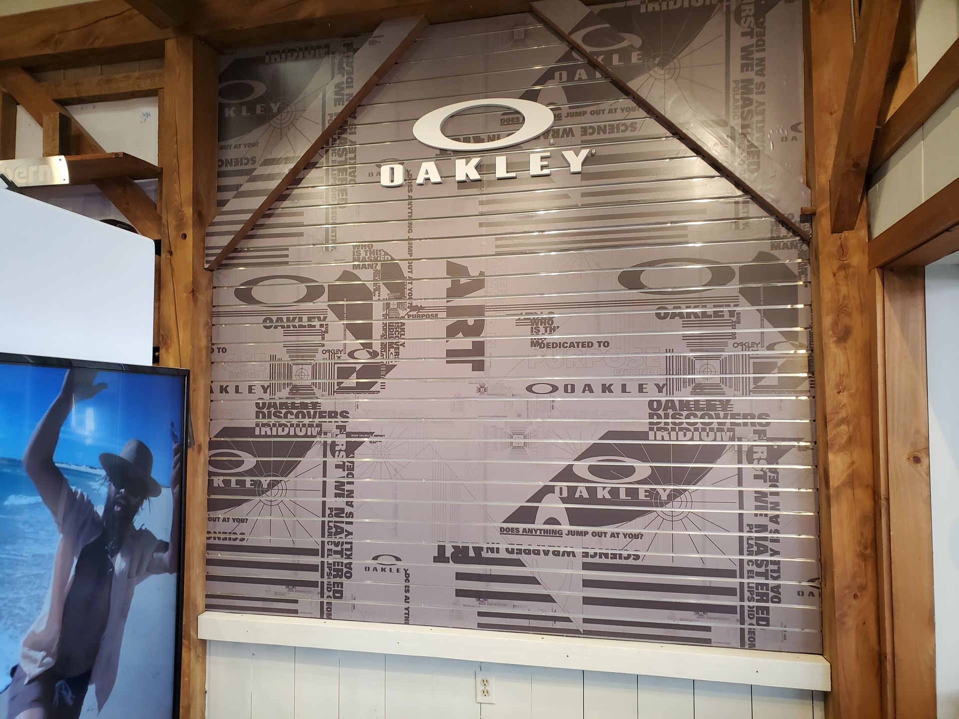 Oakley logo and design on a wall, framed by wooden beams. A display shows a person with a hat and sunglasses.