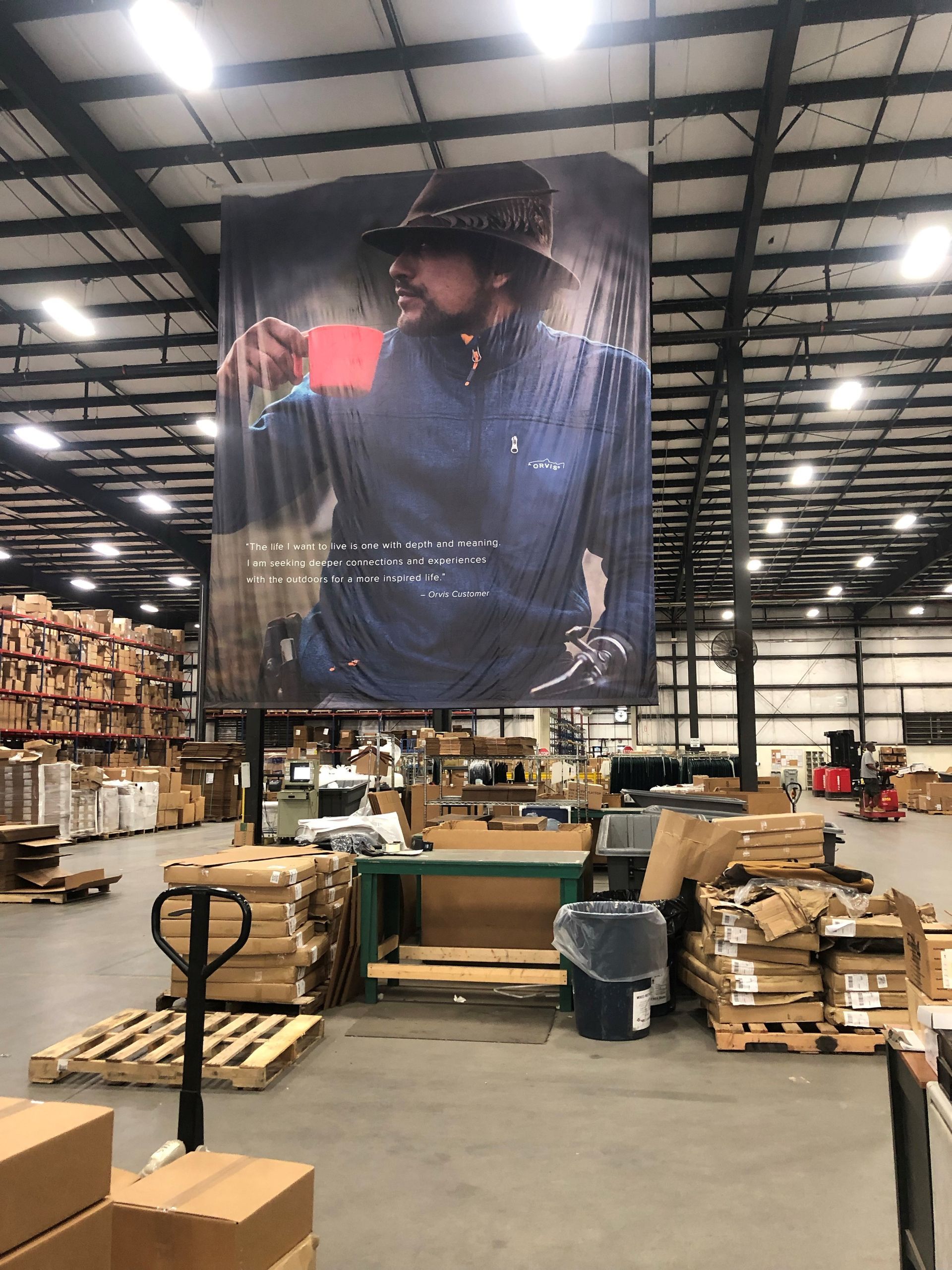 Warehouse interior with stacked pallets and a large hanging promotional banner above worktables.