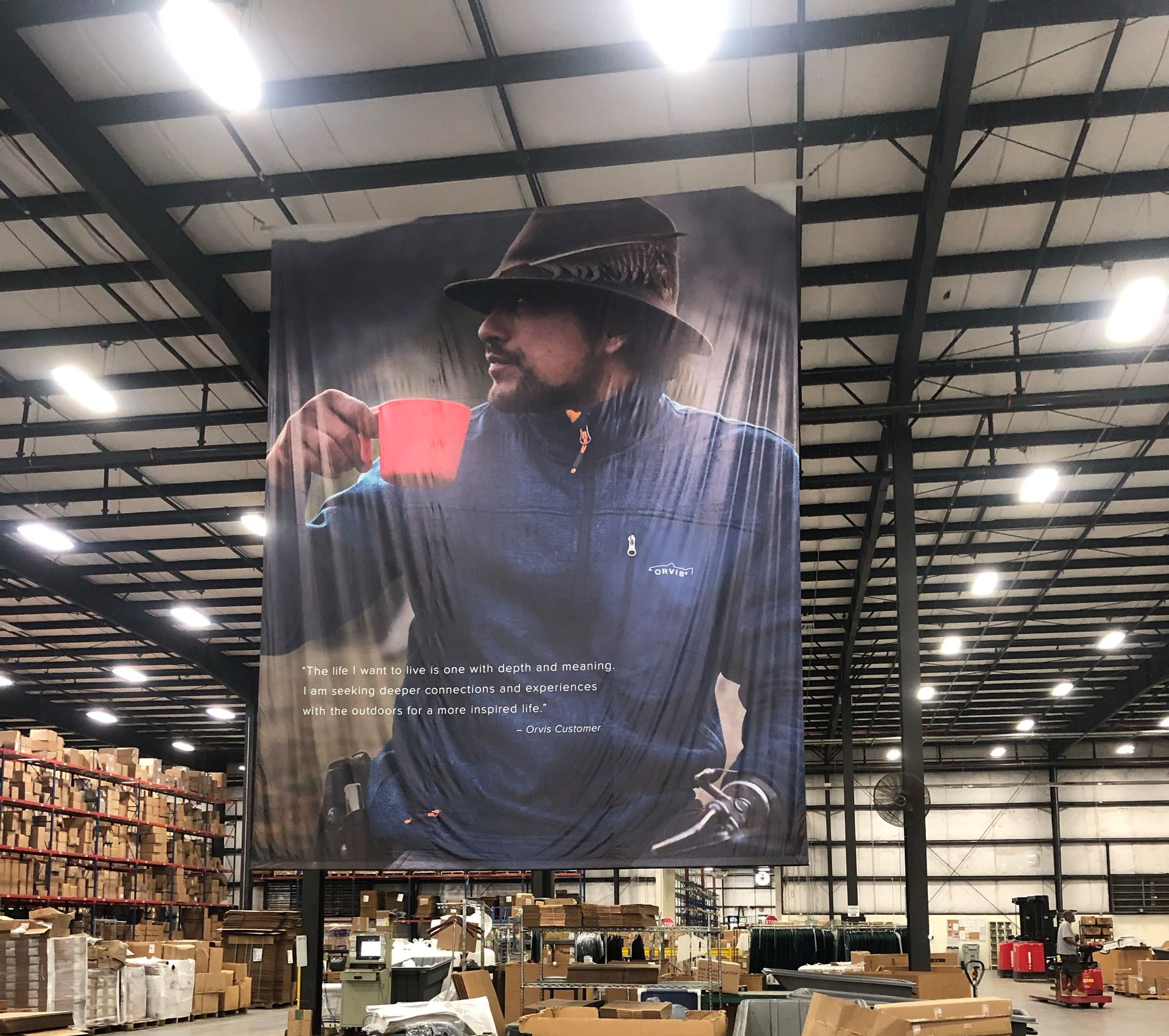 Large banner ad in a warehouse: man in hat holding a mug. Surrounded by boxes and metal beams.