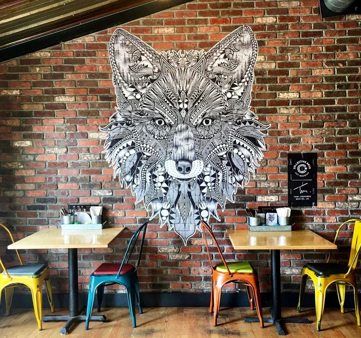 A fox mural on a brick wall above cafe tables and colorful chairs.