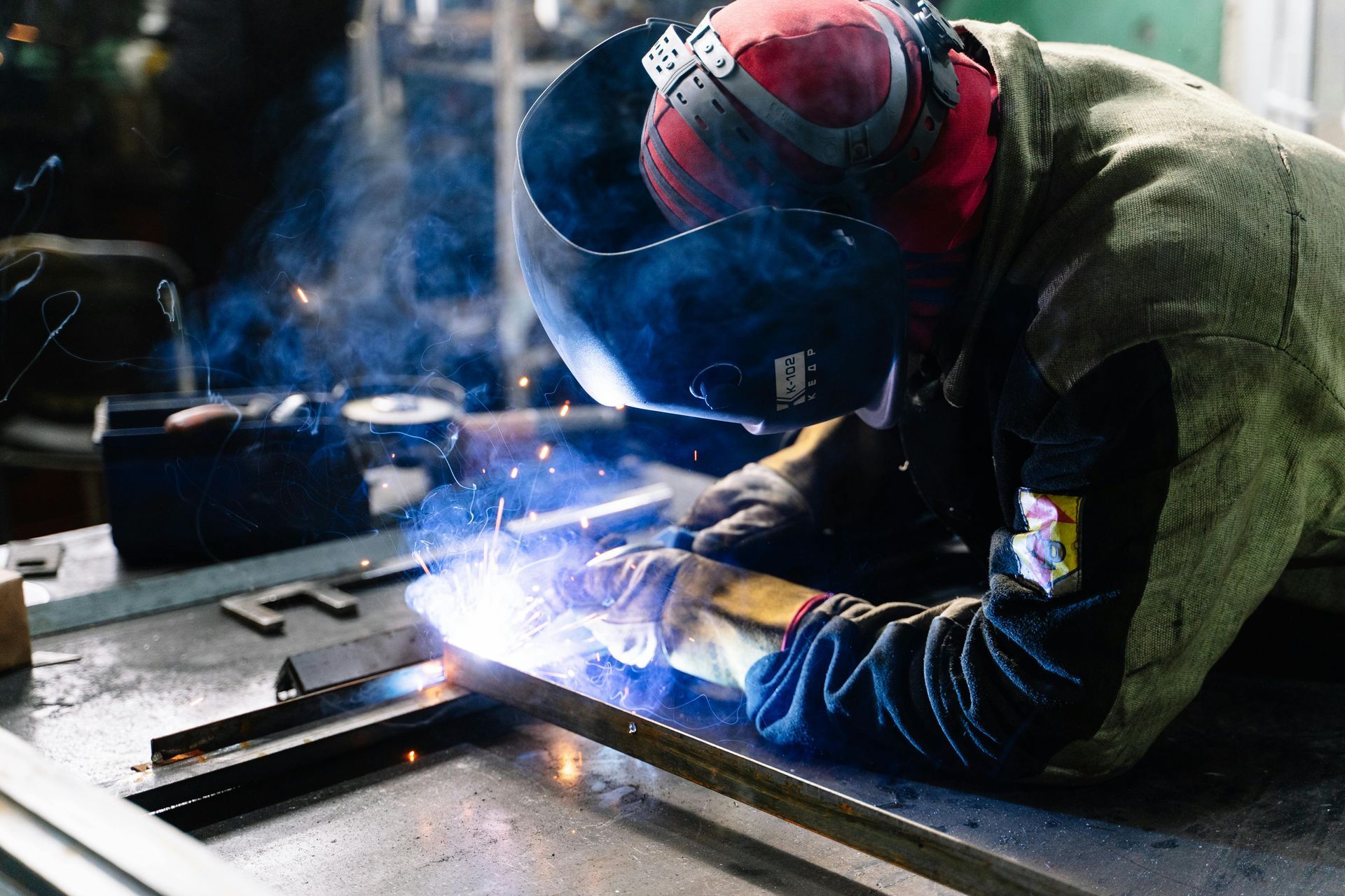 Welder in a protective helmet and gear, working with sparks on metal in a workshop.