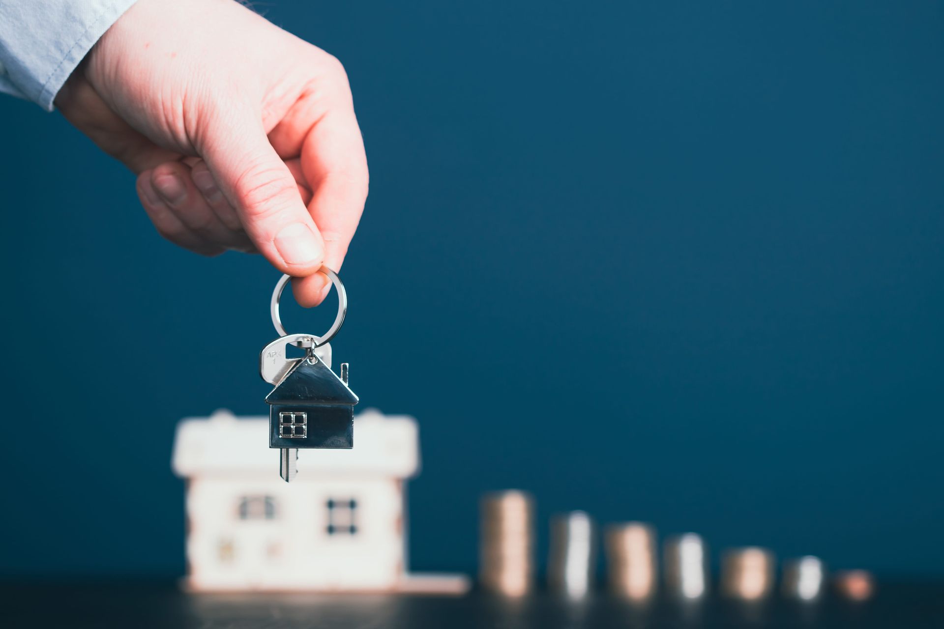 Hand holding house key above a small model house with coins on a blue background