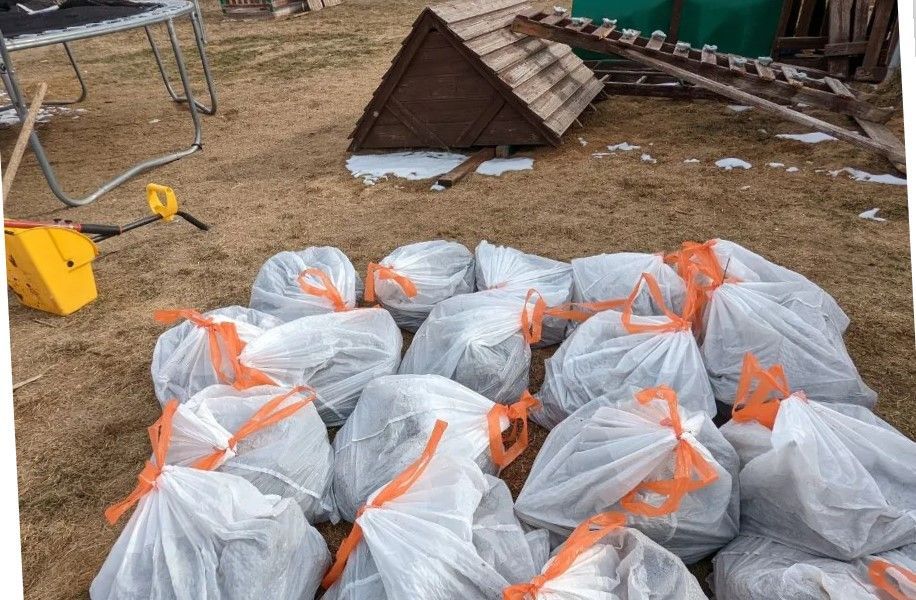 Pile of white plastic bags tied with orange ties, on brown ground, with a small wooden structure in the background.