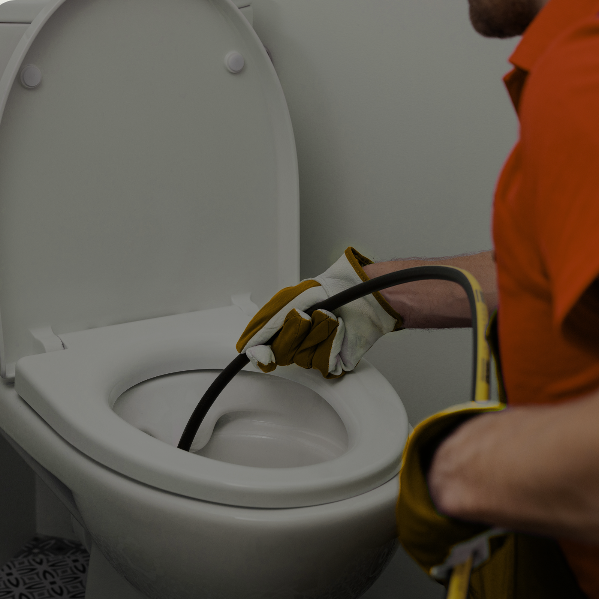 Pessoa vestindo camisa e luvas laranja usando uma serpentina de encanamento para desentupir um vaso sanitário branco em um banheiro.