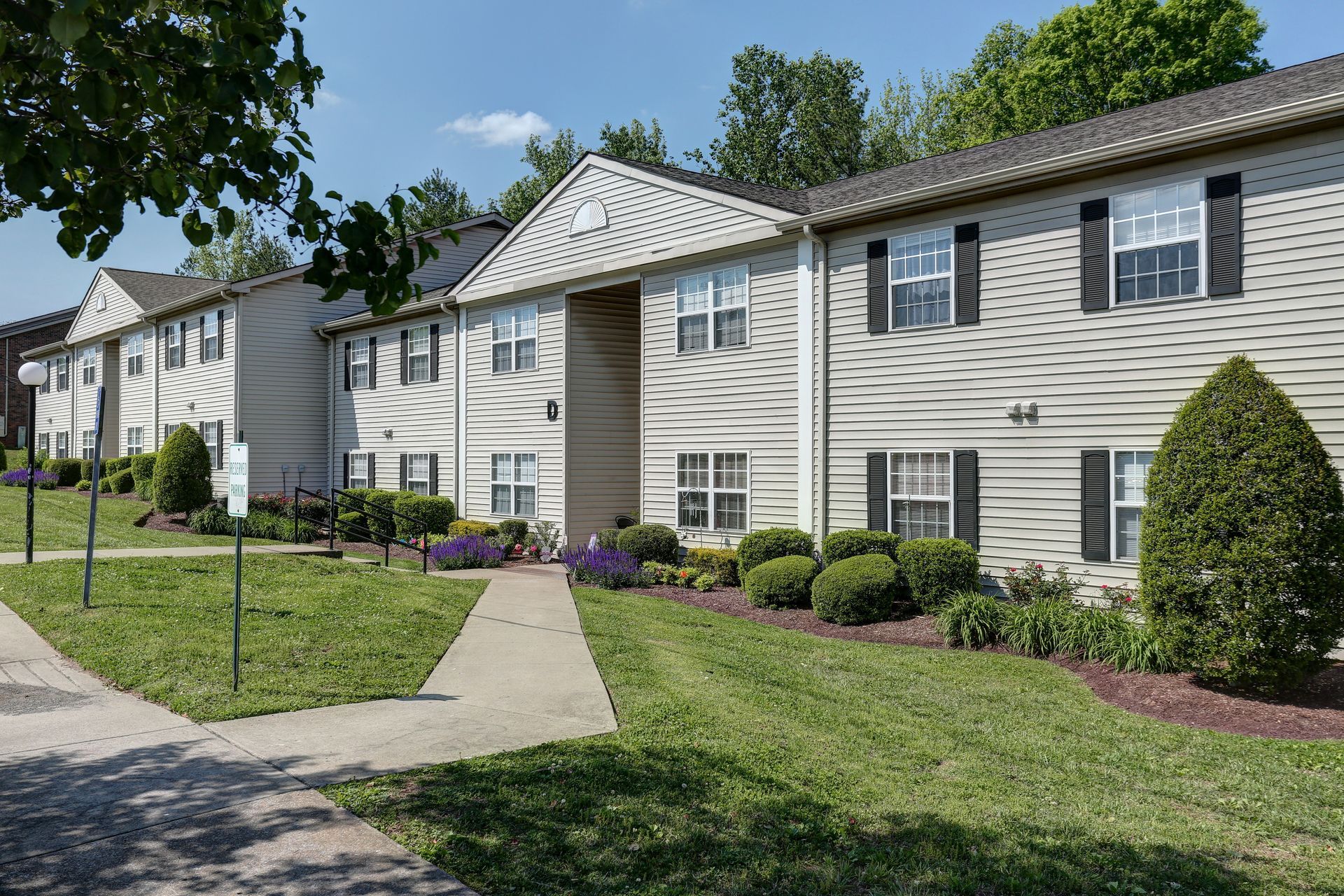 Apartment complex with beige siding, a walkway, green lawn, and trees on a sunny day