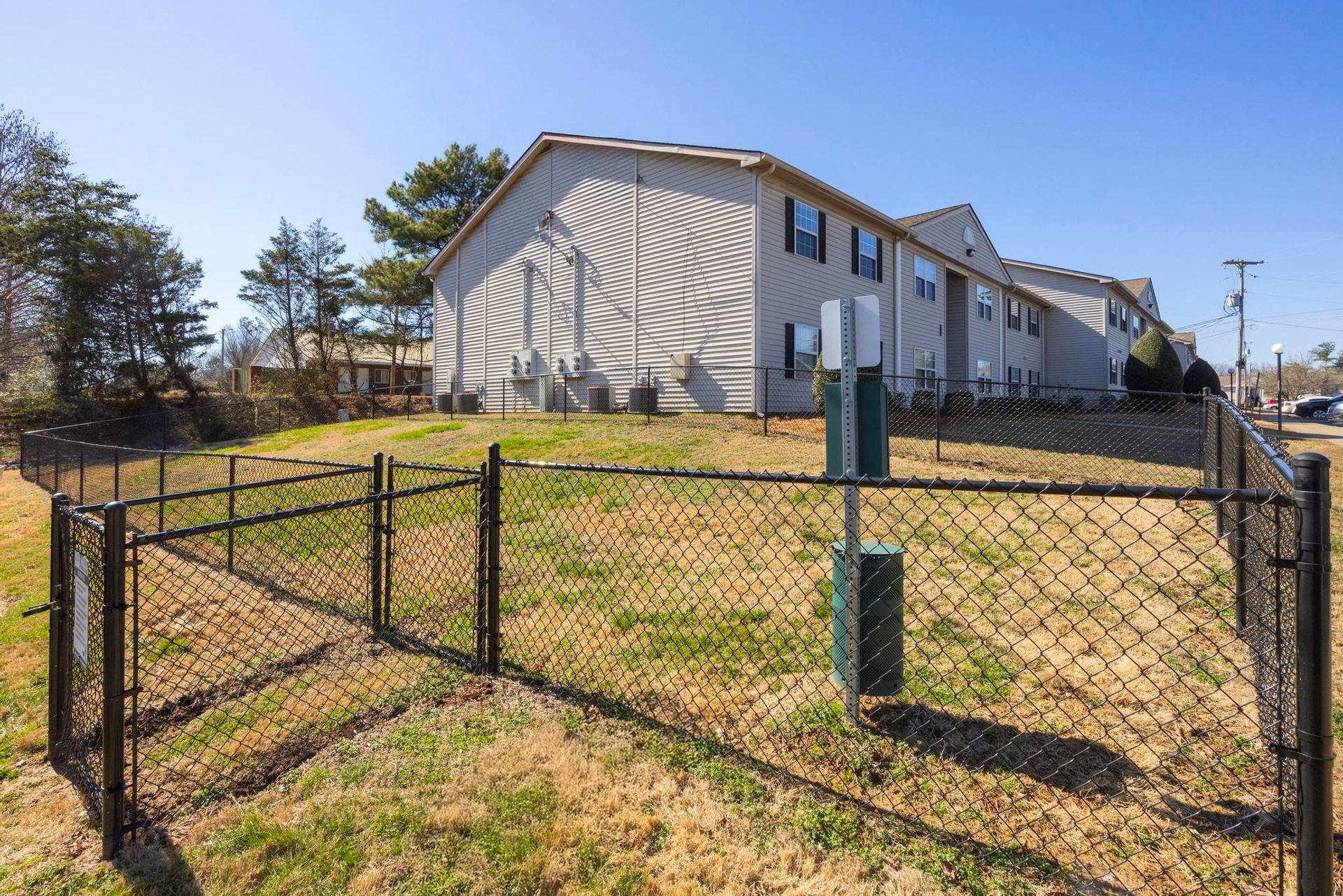 Fenced grassy lot beside a beige apartment building under a clear blue sky