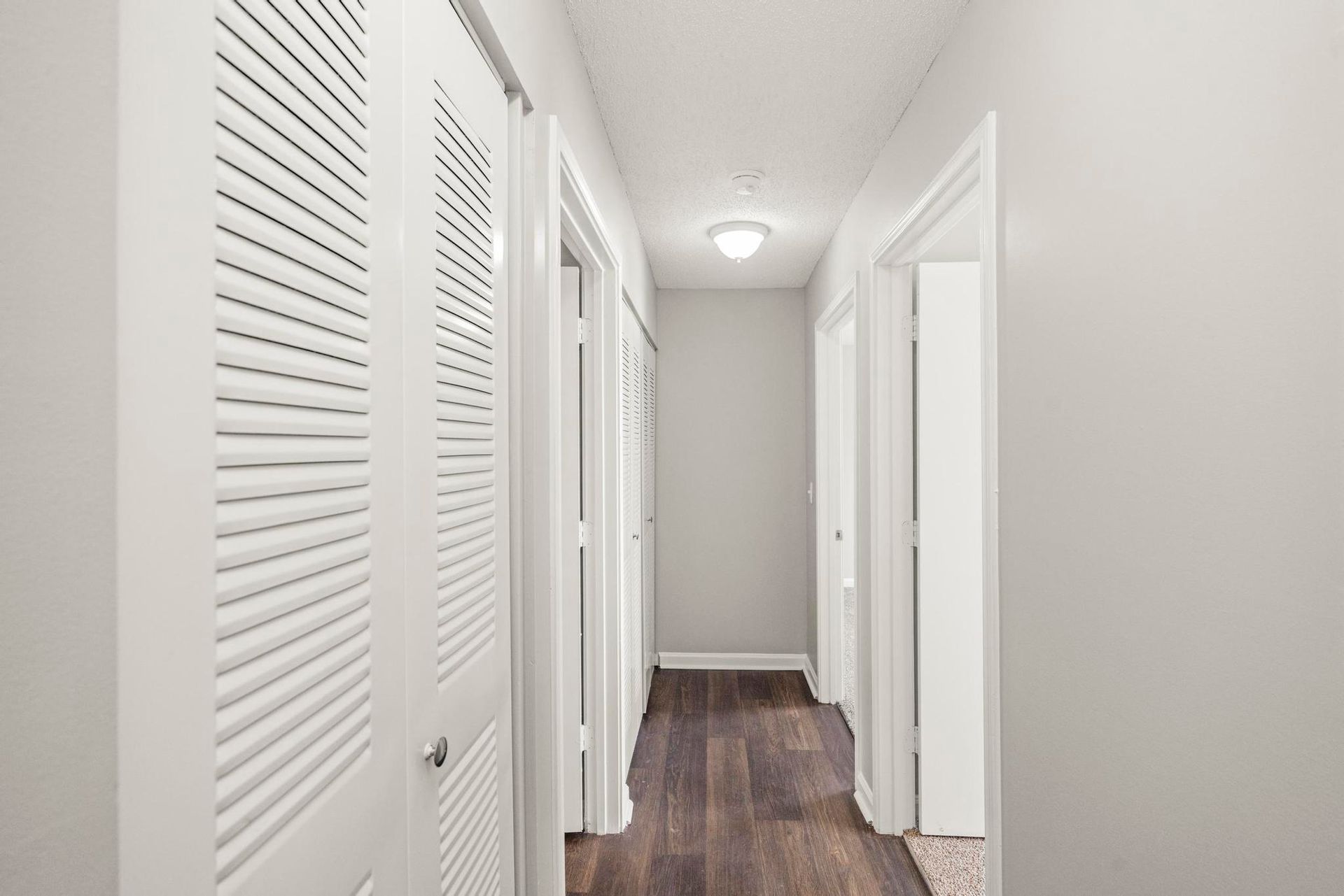 Bright hallway with white walls, louvered closet doors, and dark wood flooring leading to open doorways