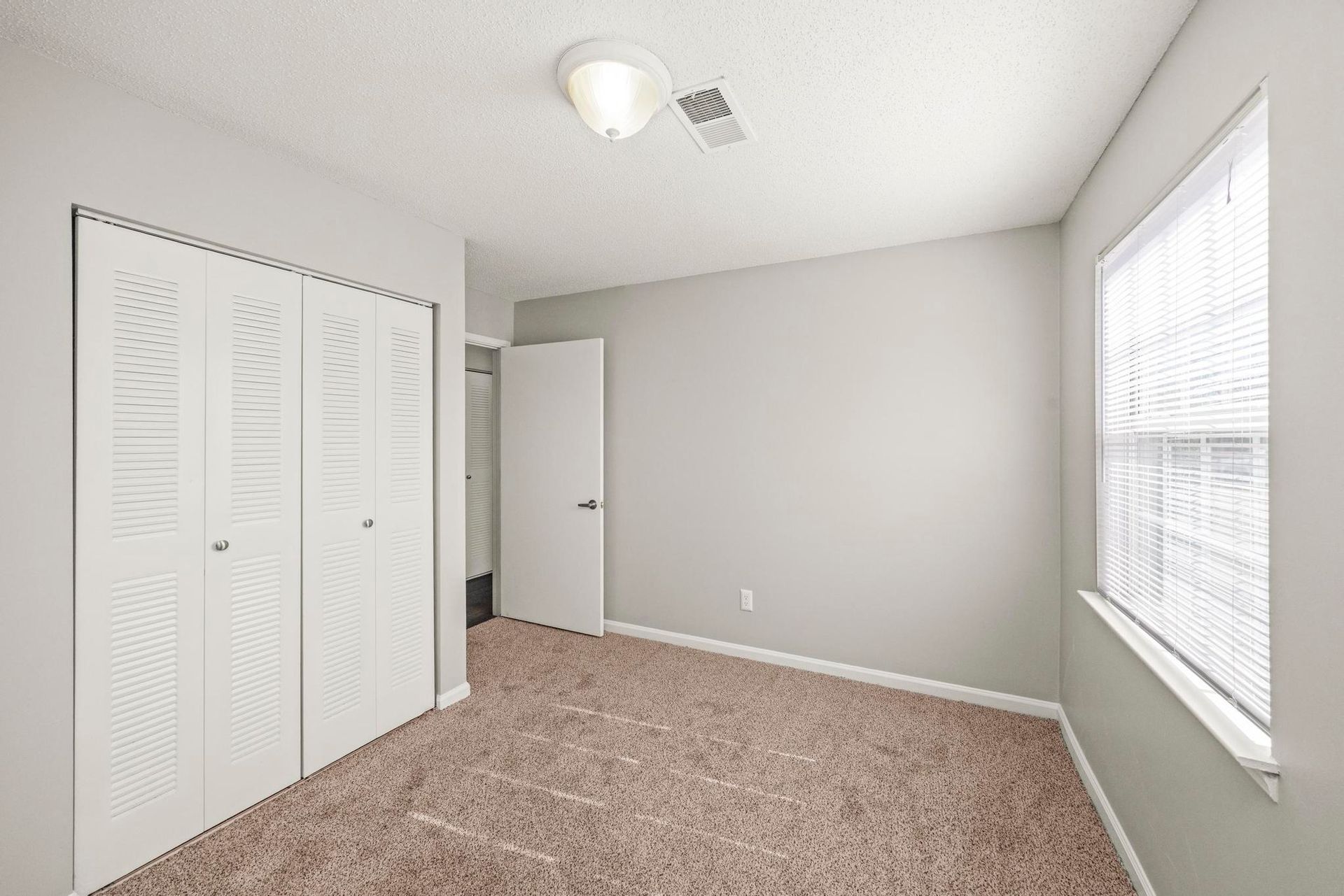 Empty beige bedroom with carpet, closet doors, a window, and an open doorway.