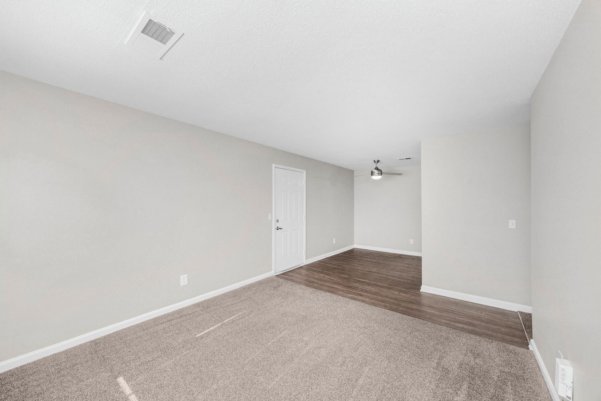 Empty carpeted living room with gray walls, white door, and wood flooring in back area