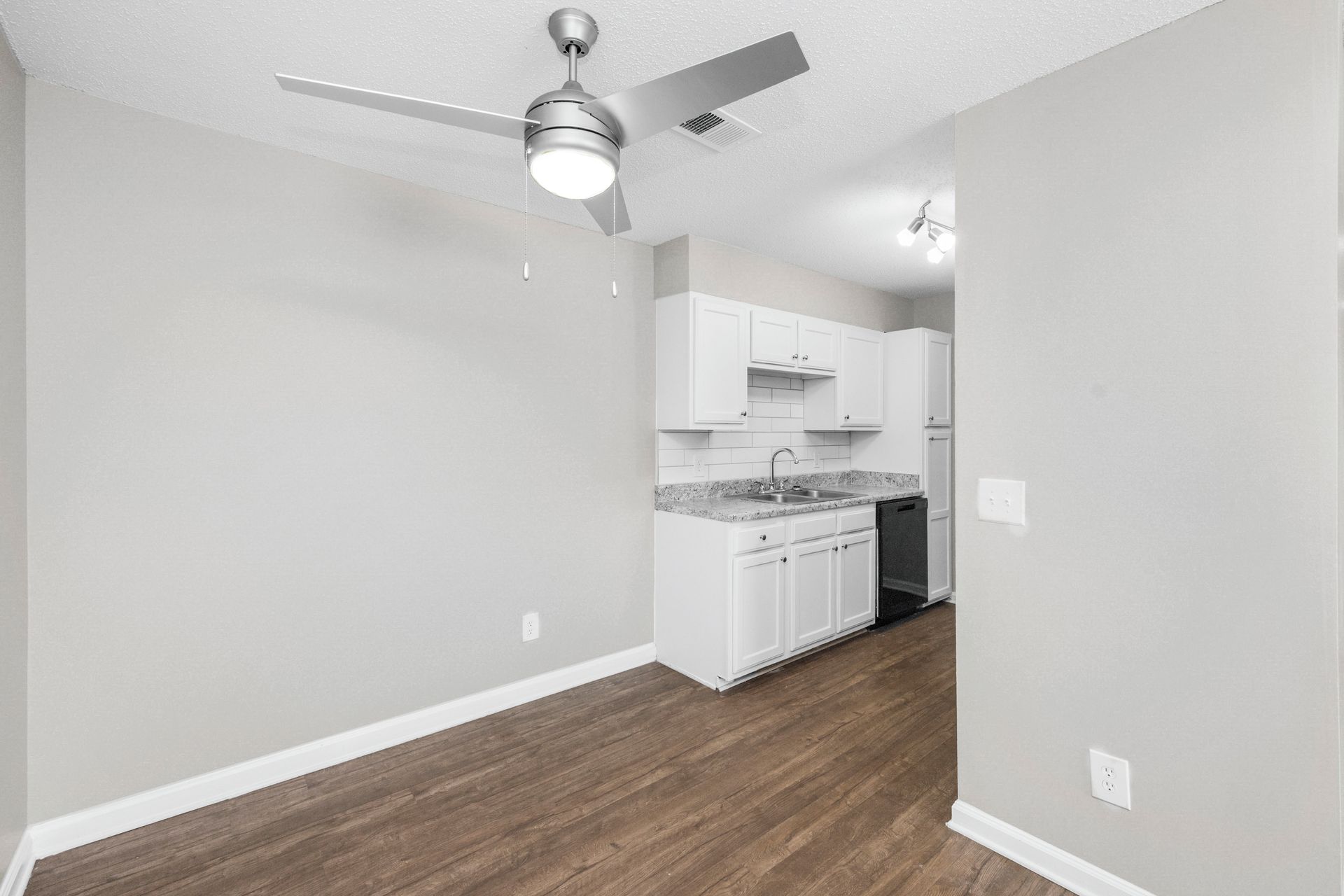 Empty room with gray walls, wood flooring, ceiling fan, and a small white kitchen area.