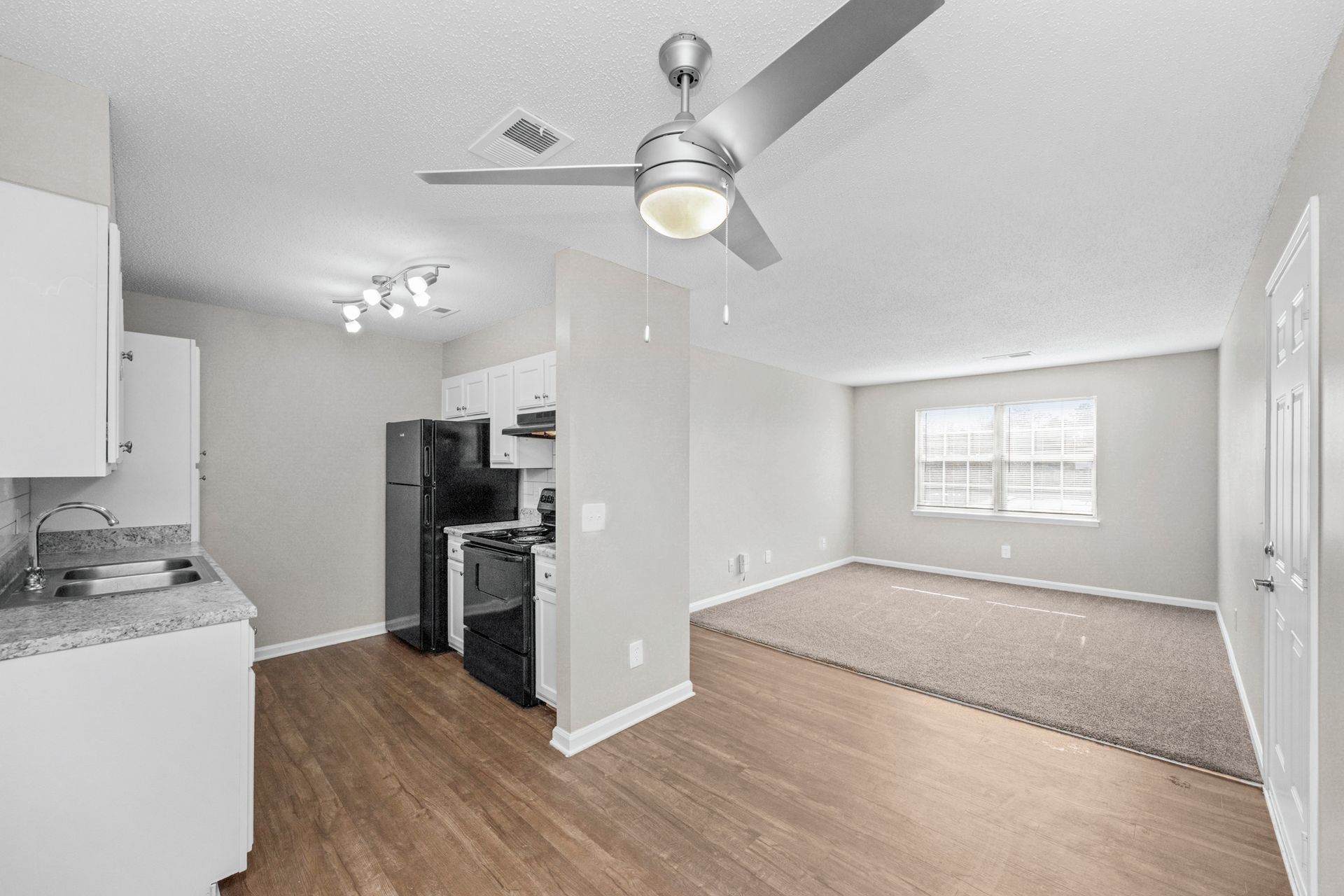 Open-concept kitchen and living room with wood floors, ceiling fan, stainless appliances, and carpeted sitting area.