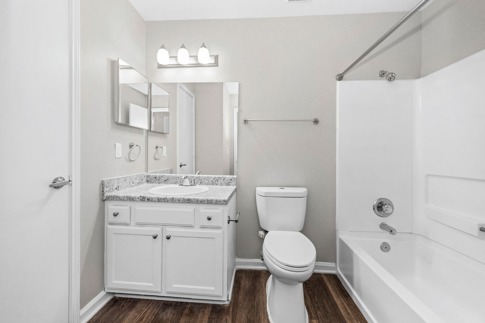 Bright white bathroom with vanity, toilet, and shower-tub combo, dark wood floor, and three-light fixture.