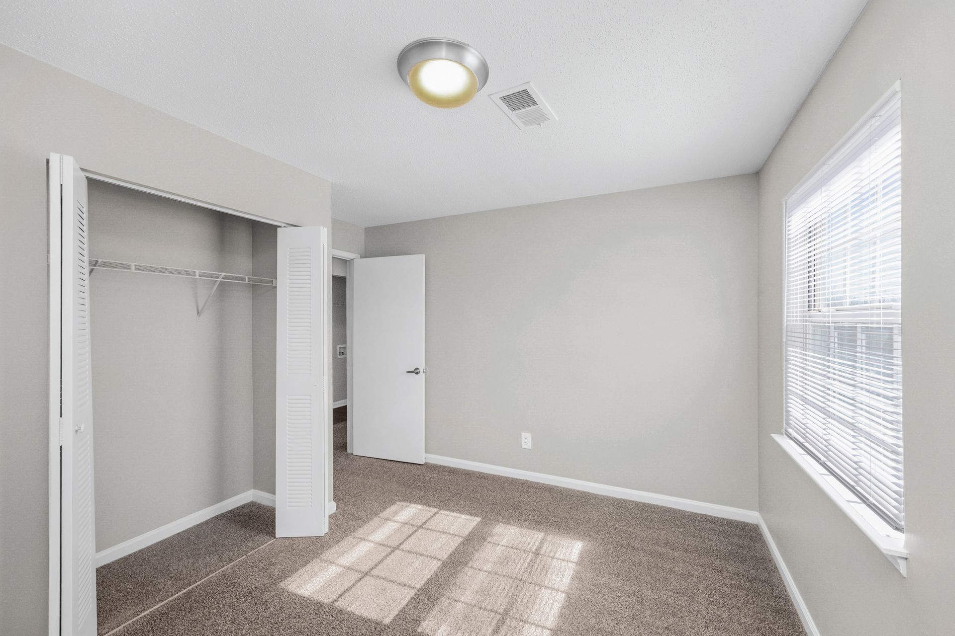 Empty beige bedroom with carpet, open closet, white door, and window blinds letting in sunlight
