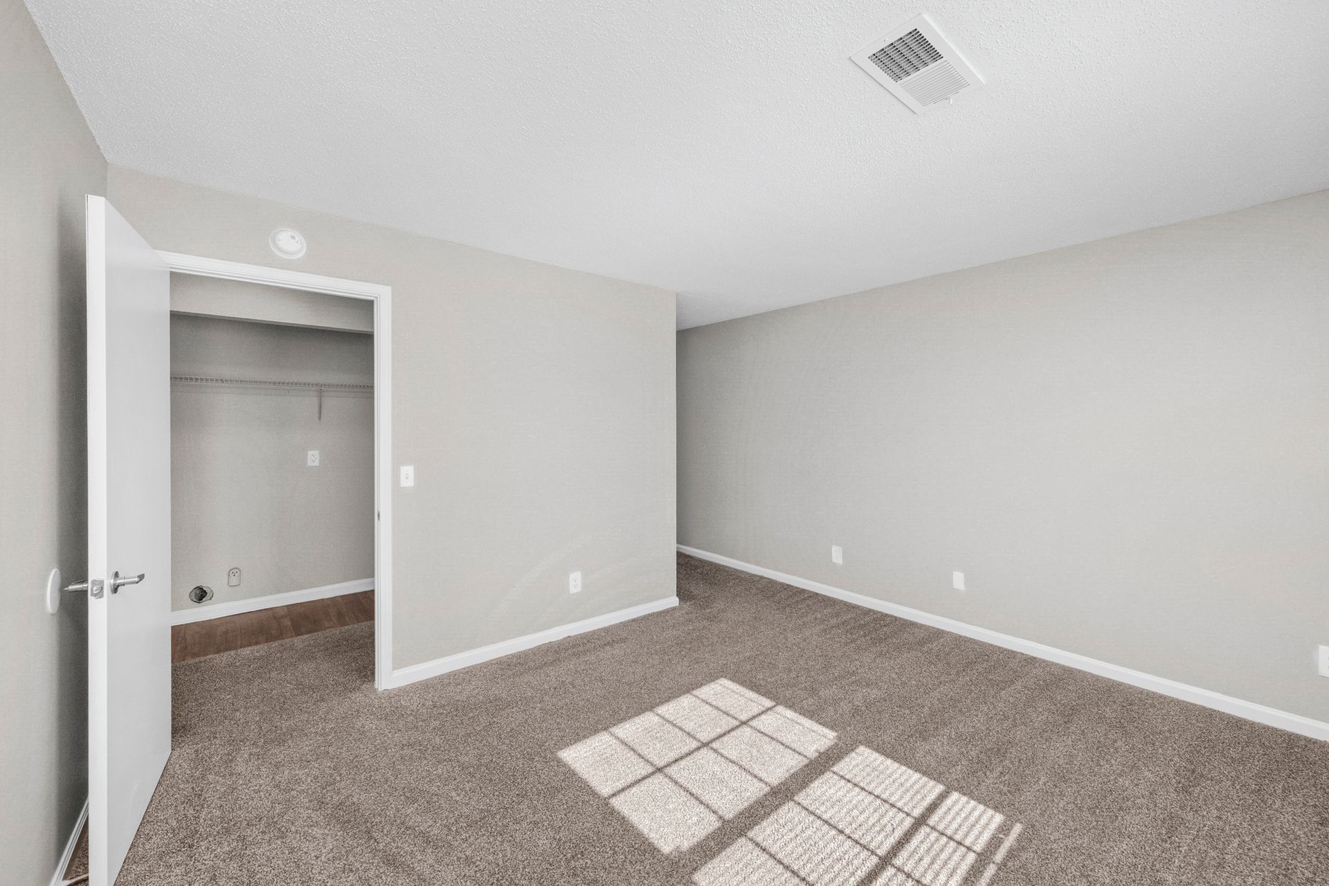 Empty beige bedroom with carpet, open closet, and sunlight on the floor