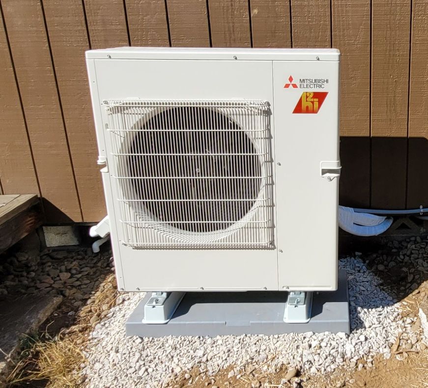 Heavy Metal HVAC llc - Mitsubishi heat pump unit, outdoors, mounted on concrete base, next to a wooden wall.