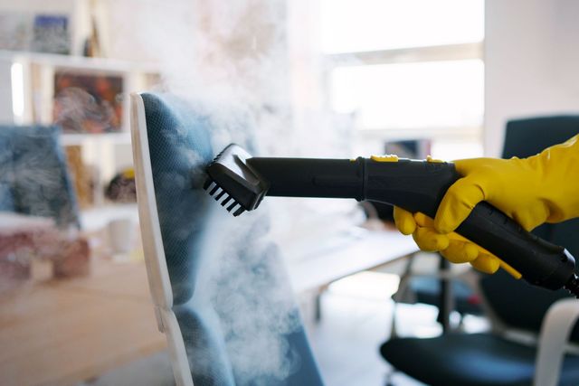 Handheld steam cleaner spraying a glass surface, with a gloved hand in the foreground
