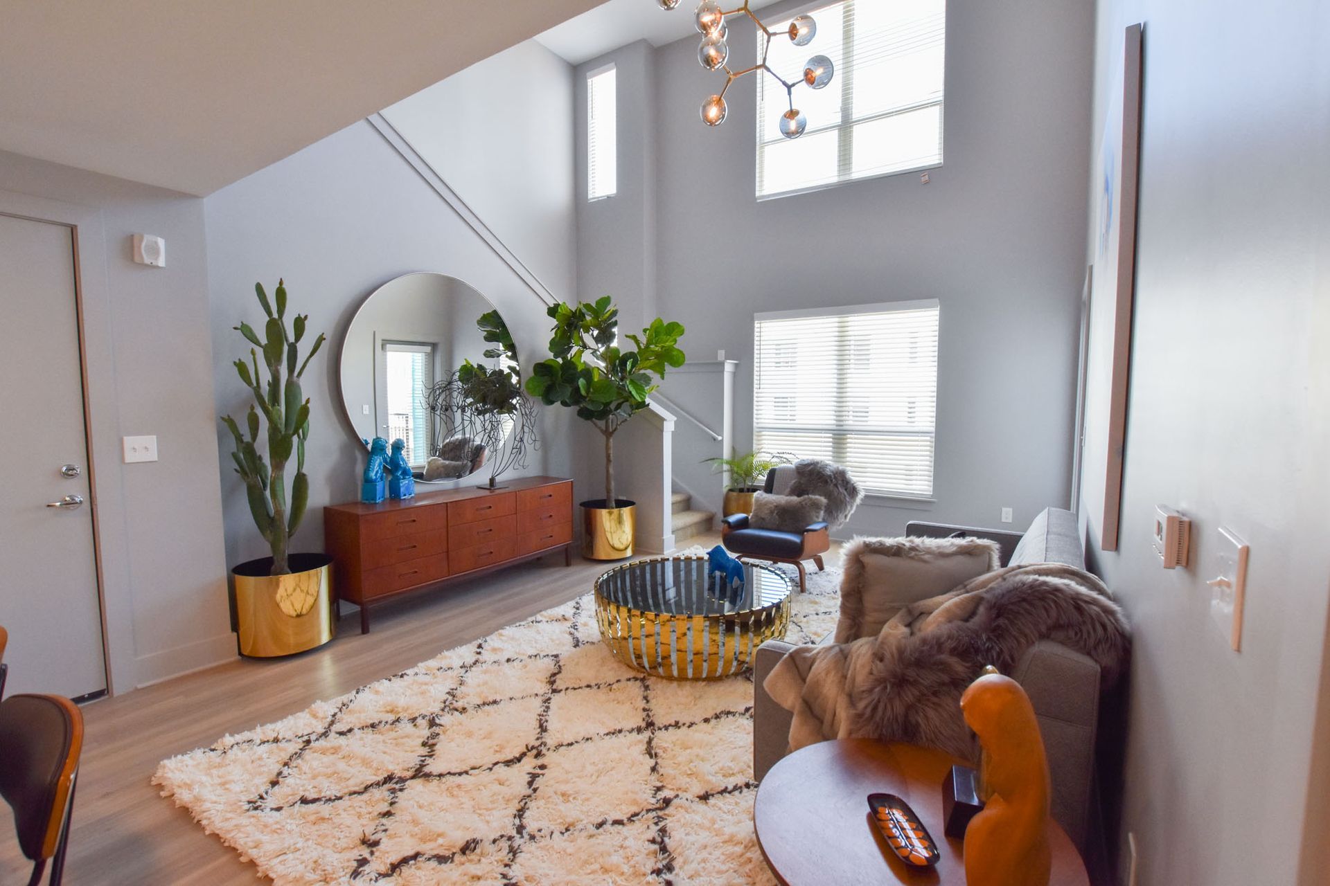 Bright living room with sofa, round mirror, plants, and patterned rug beneath a high ceiling.
