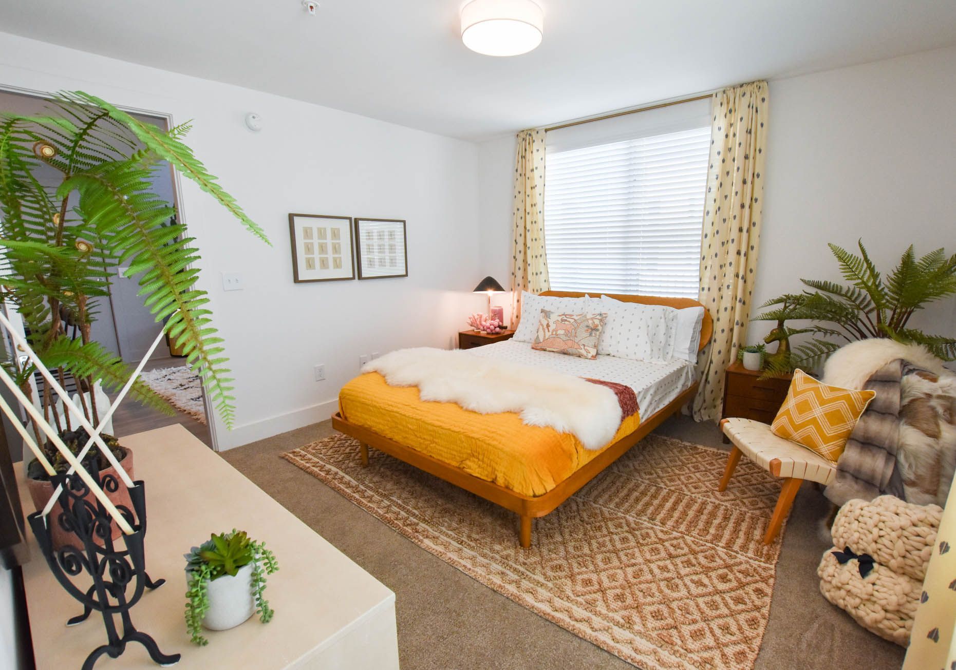 Bright bedroom with a mustard bed, patterned rugs, plants, and a window with white blinds