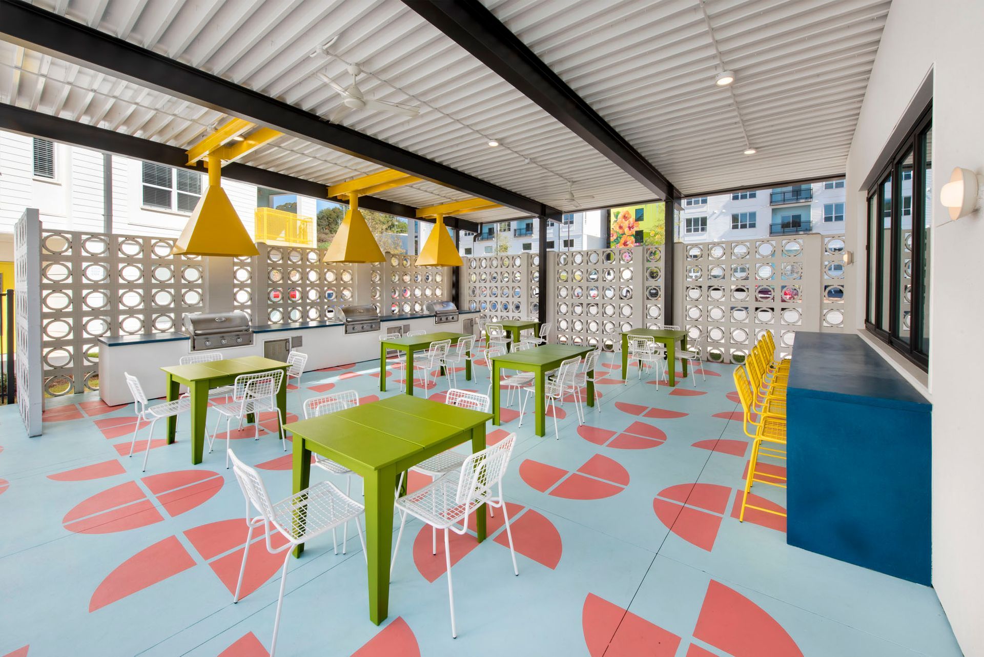 Colorful open-air café with green tables, white chairs, yellow pendant lights, and patterned floor tiles