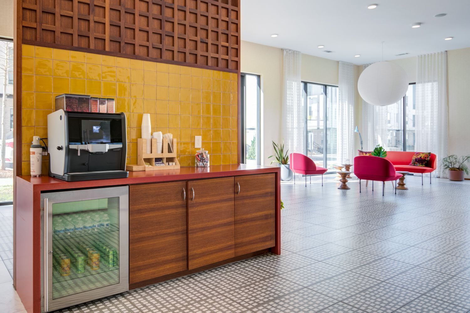 Modern lounge with coffee station, red chairs, and large windows overlooking a bright tiled floor.