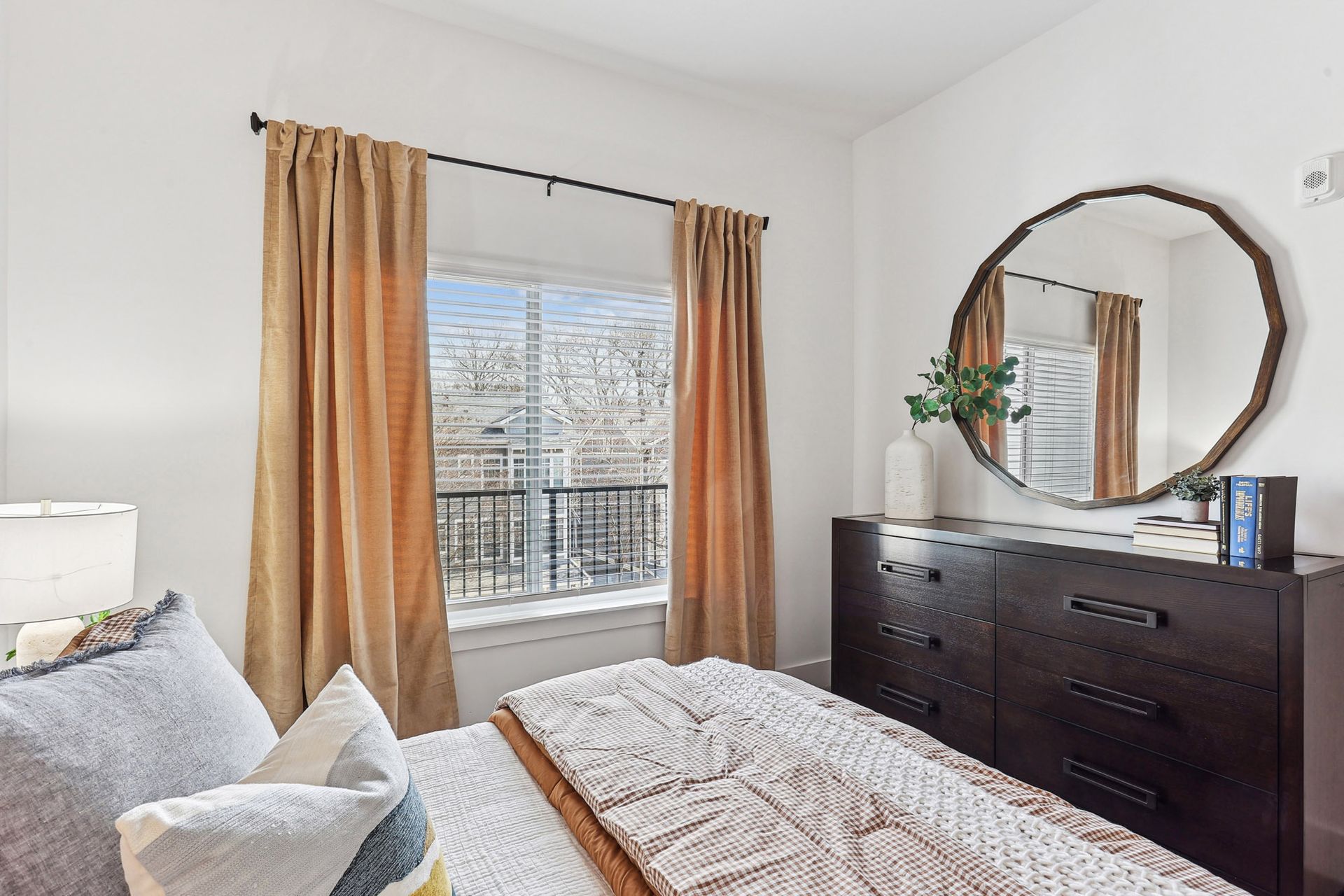 Bright bedroom with a bed, dresser, mirror, and window with tan curtains overlooking a balcony