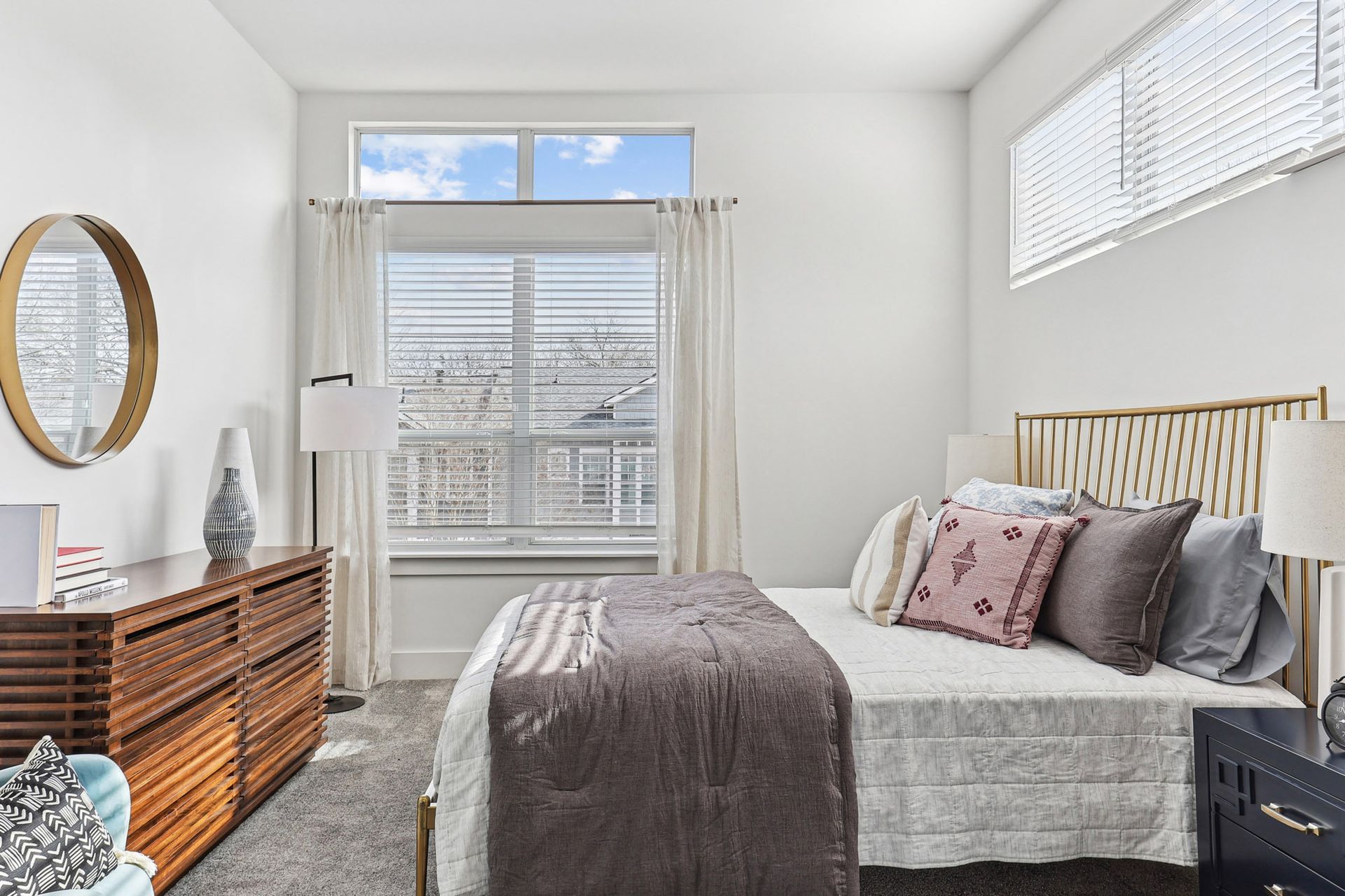 Bright bedroom with a made bed, dresser, mirror, and large windows with blinds and curtains