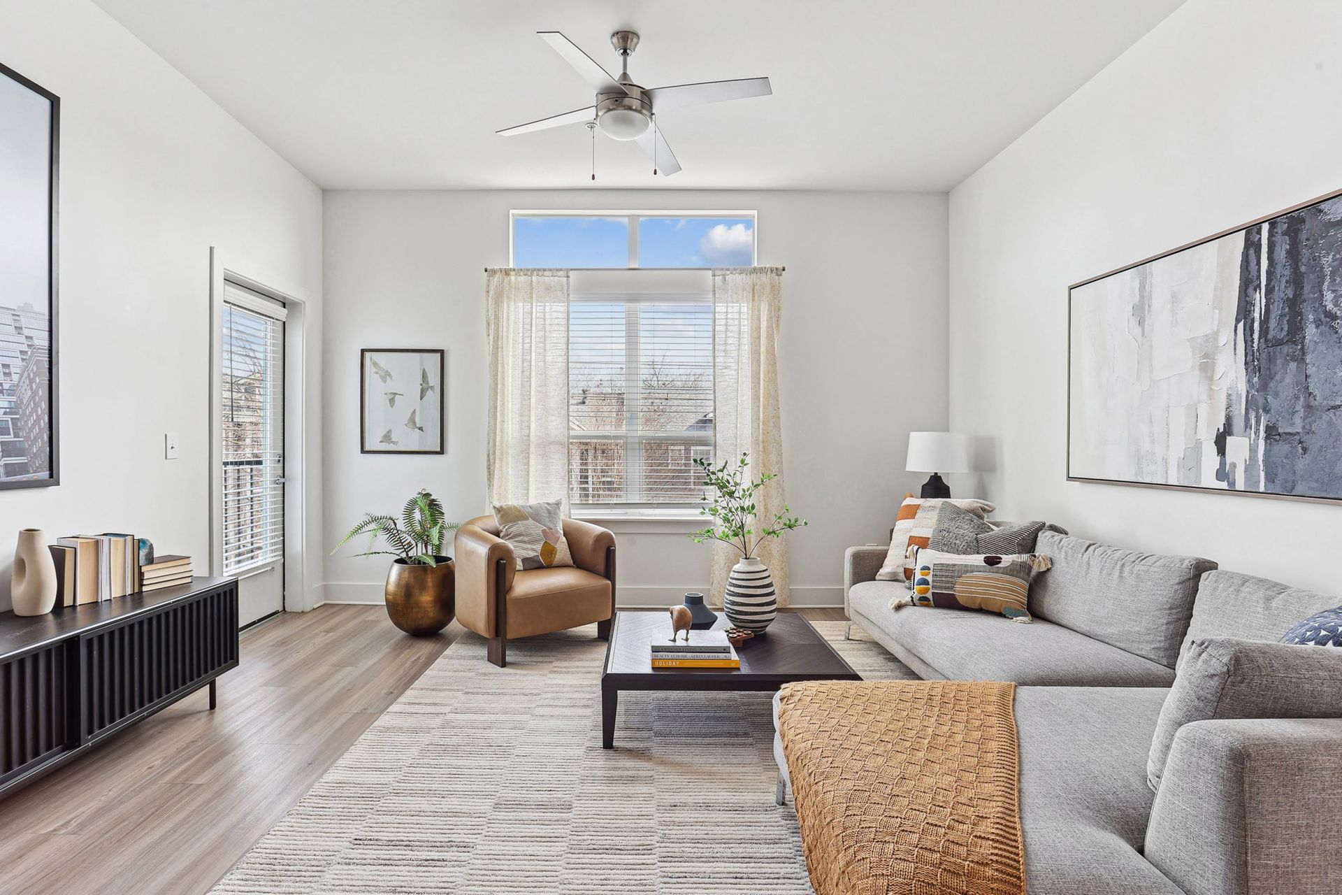 Bright modern living room with gray sofa, armchairs, rug, plants, and large window with sheer curtains