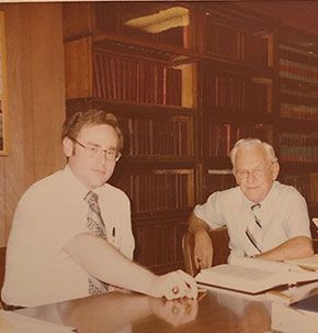 Two men are sitting at a table in front of a bookshelf.