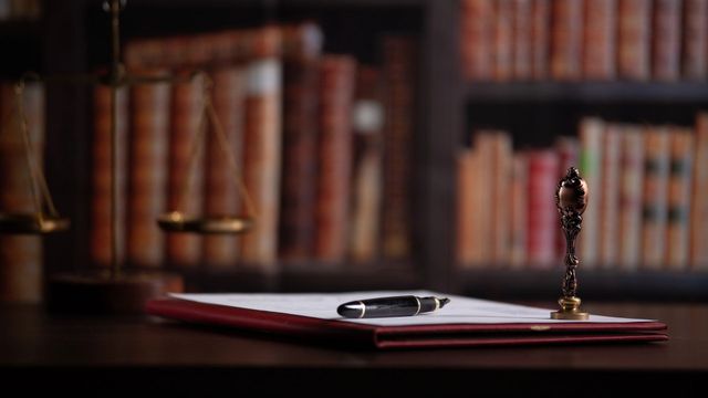 Scrivania con occhiali e penna su una cartella rossa, con scaffali sullo sfondo.