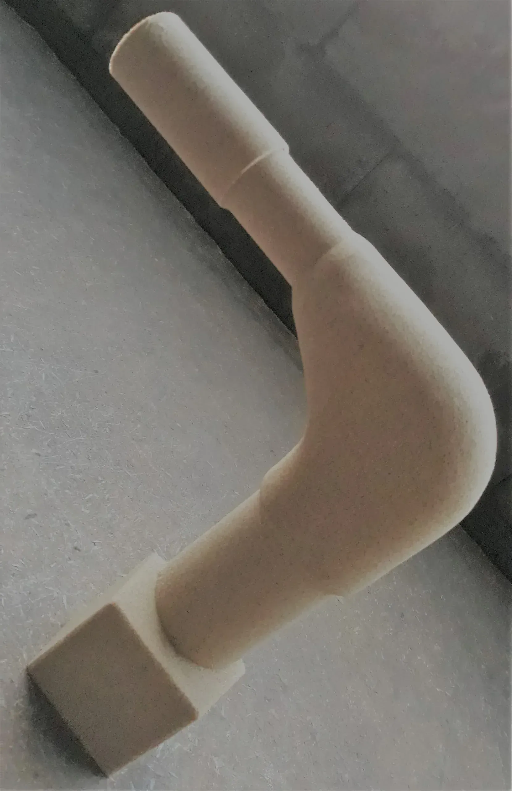 Pipa in ceramica beige, a forma di L con base quadrata e parte superiore angolata, su una superficie grigio chiaro.