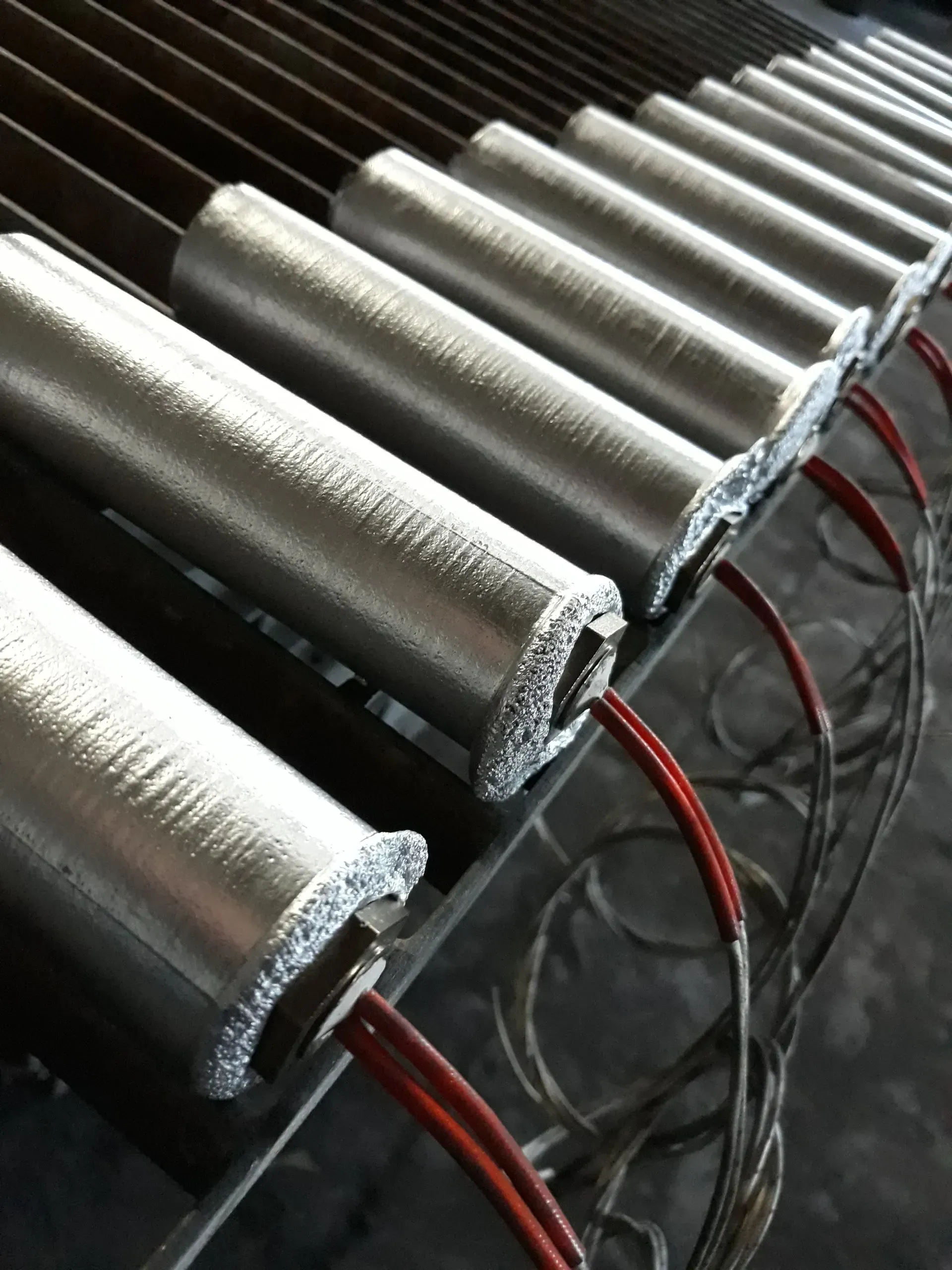 Primo piano di una fila di elementi riscaldanti industriali cilindrici argentati con fili rossi esposti.