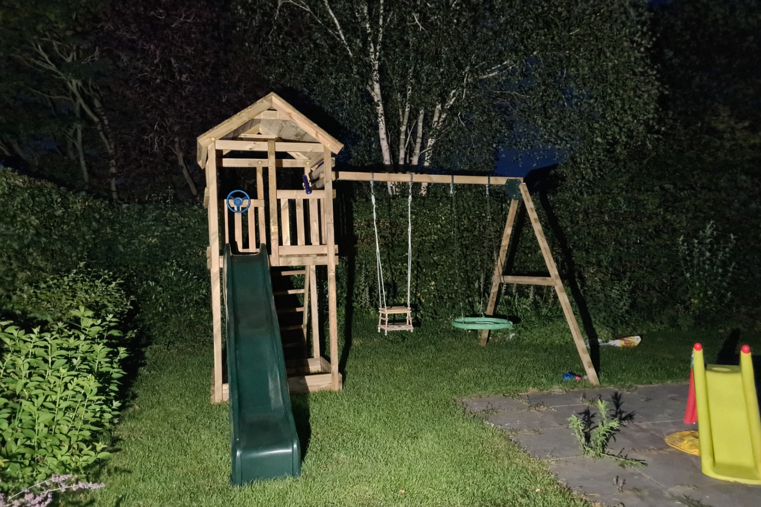 Houten speeltoestel met een glijbaan, schommels en een kleine, gele glijbaan op een gazon.