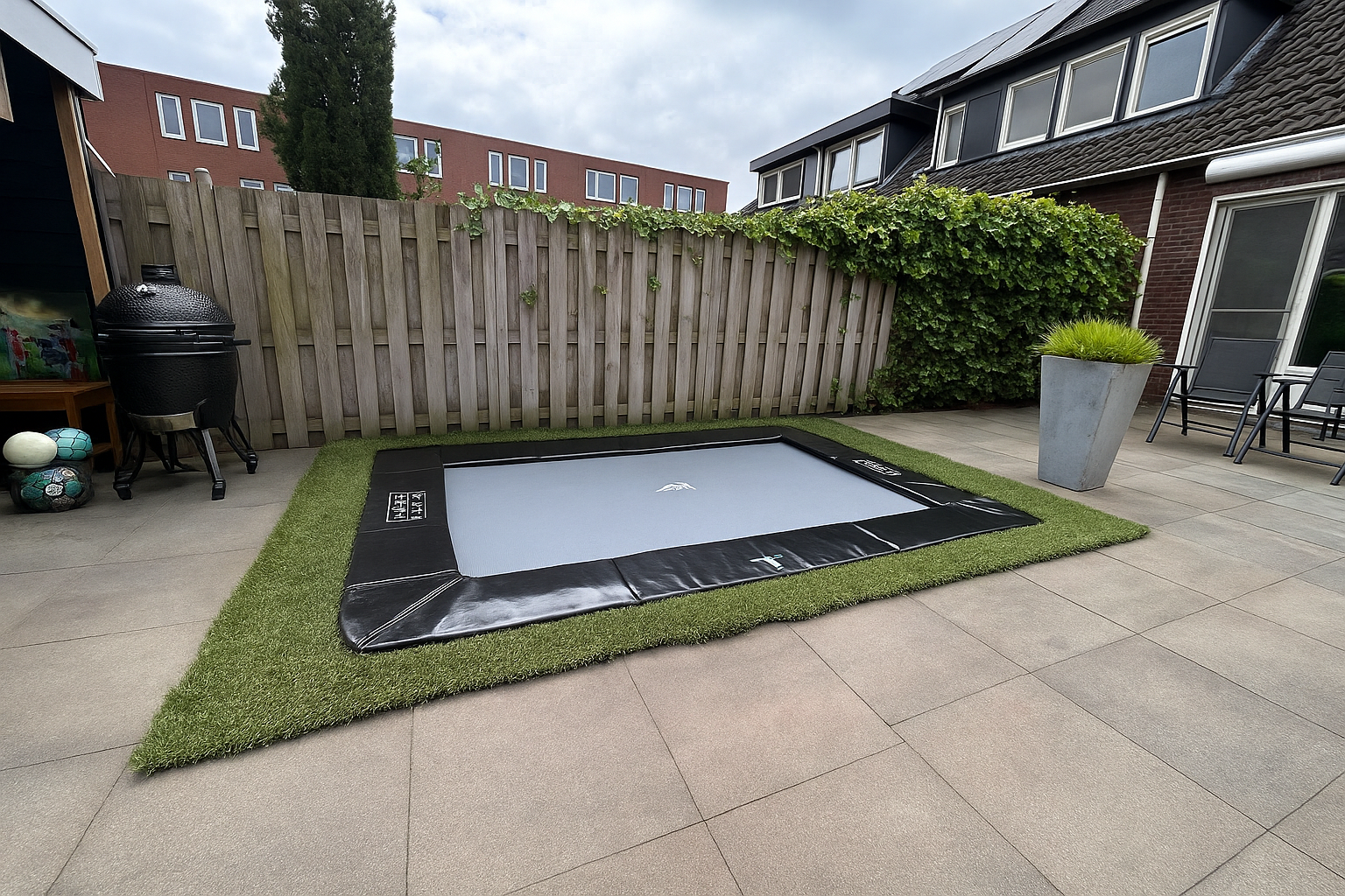 Een rechthoekige trampoline, ingegraven op kunstgras, in een betegelde achtertuin, met een houten schutting en een huis op de achtergrond.