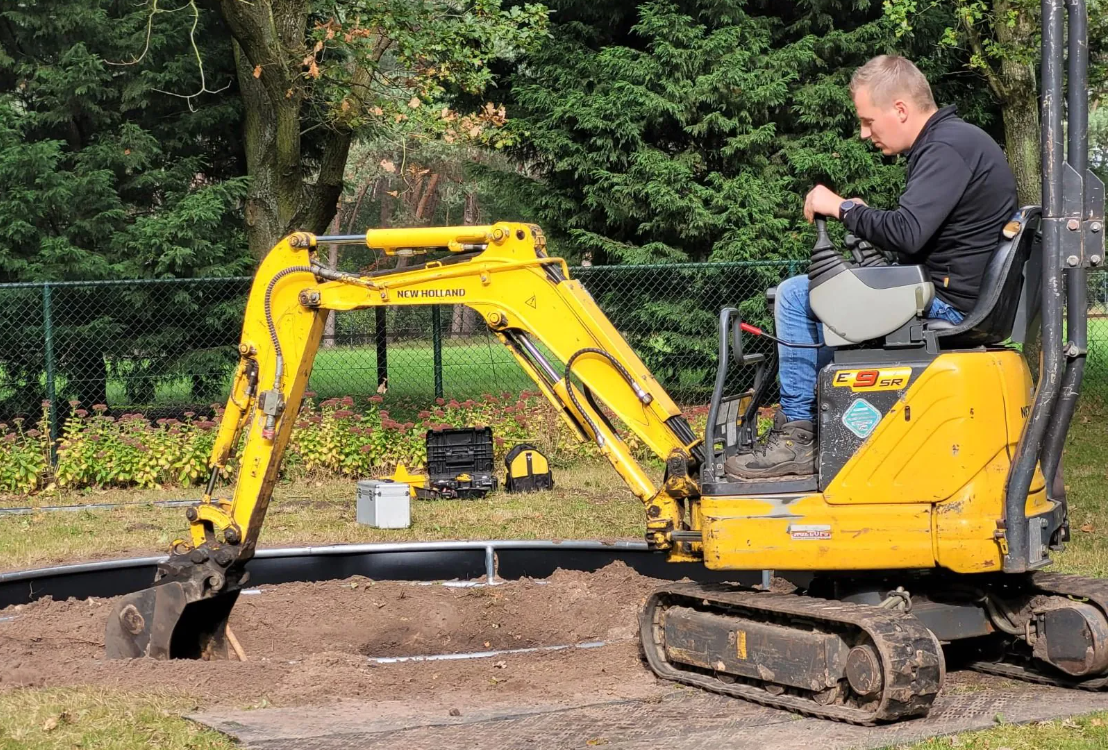 Een man bedient een gele minigraafmachine om een ​​gat te graven in een grasveld.