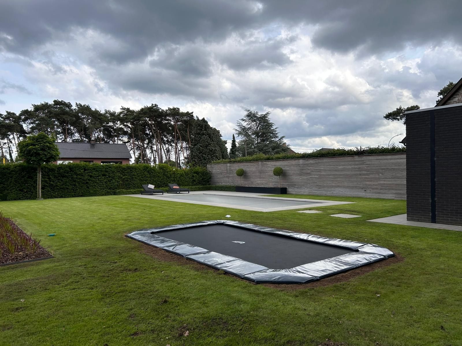 Een scène uit een achtertuin met een rechthoekige, ingegraven trampoline, een groen gazon, bomen en een bewolkte hemel.