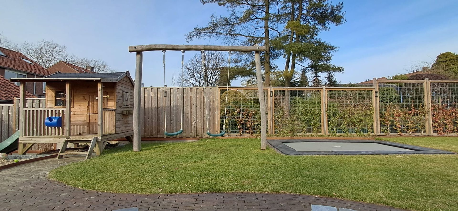 Een achtertuin met een houten speelhuisje, schommel, trampoline en een omheinde tuin op een zonnige dag.