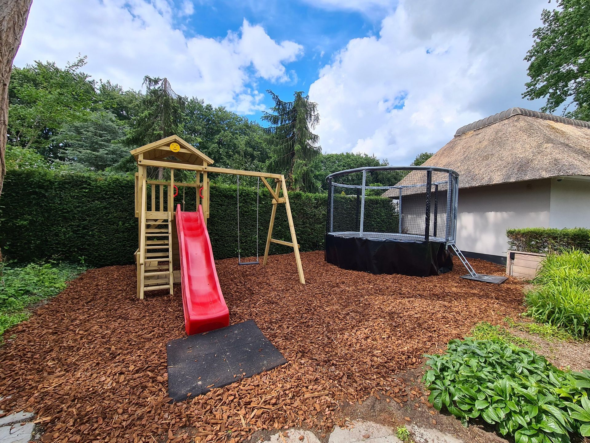 Speeltuin met schommel, glijbaan en trampoline op een ondergrond van houtsnippers, vlakbij een huis met een rieten dak.