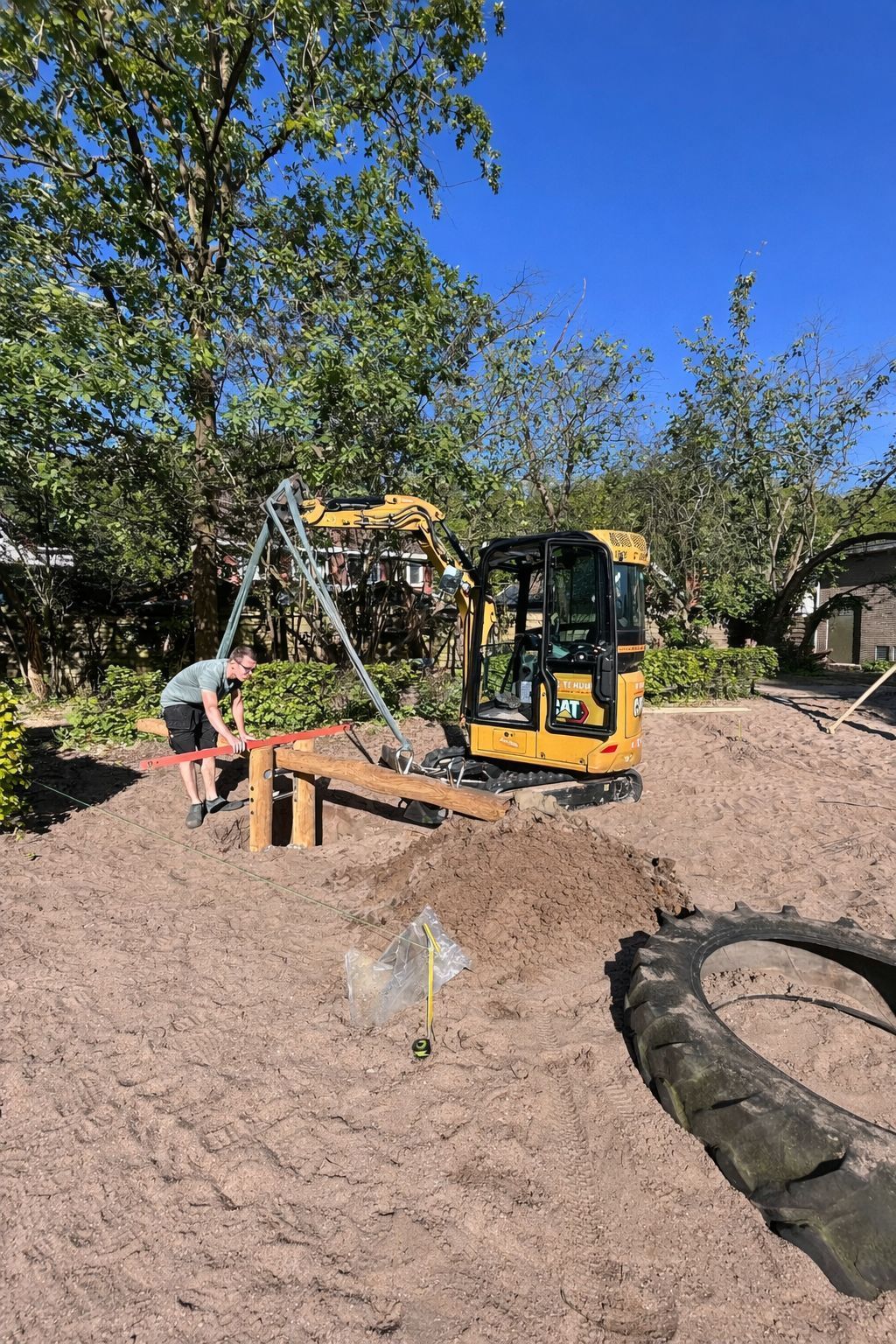 Een minigraafmachine graaft een rechthoekig gebied uit in een grasrijke achtertuin, omgeven door struiken en een huis.