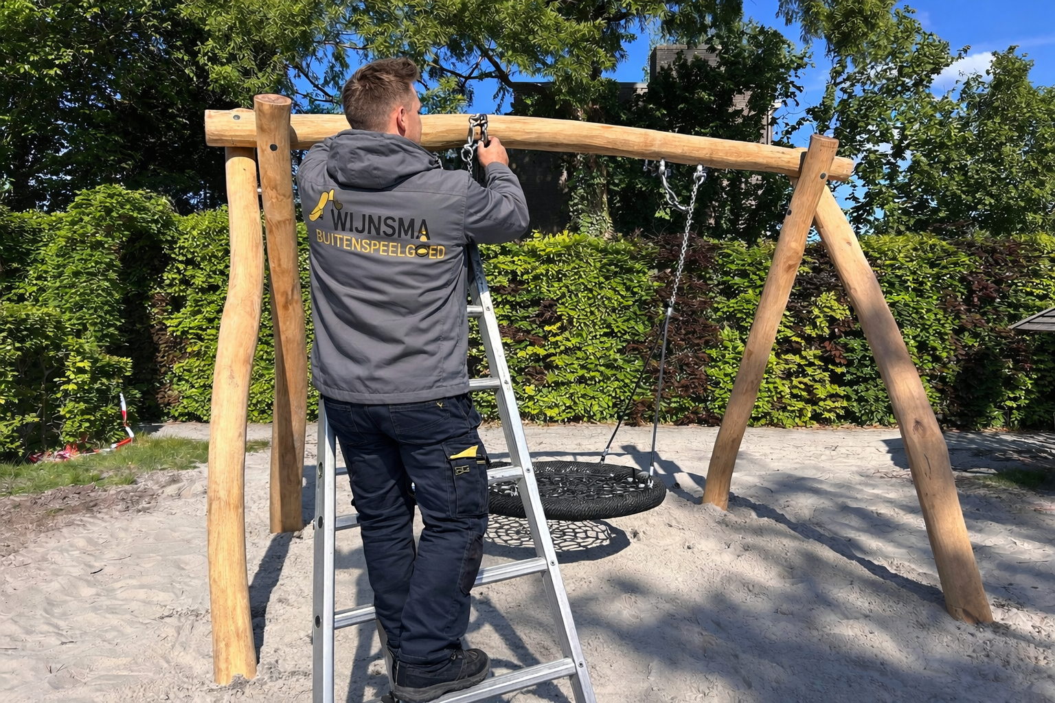 Een man staat op een ladder om een bandenschommel te bevestigen aan een houten schommelset in een zanderige speelruimte.