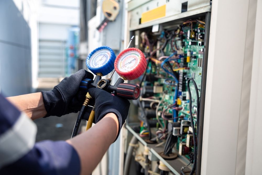 Technician using gauges to service an air conditioning unit; wires and circuits visible.