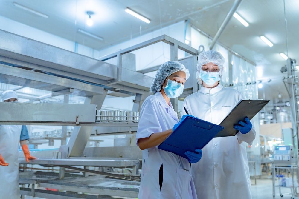 Two masked lab workers in white coats reviewing clipboard notes in a bright industrial lab