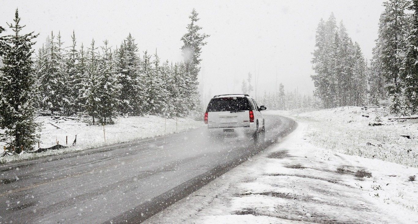 A white SUV driving along a winding, snow-covered road lined with evergreen trees during a winter snowstorm.