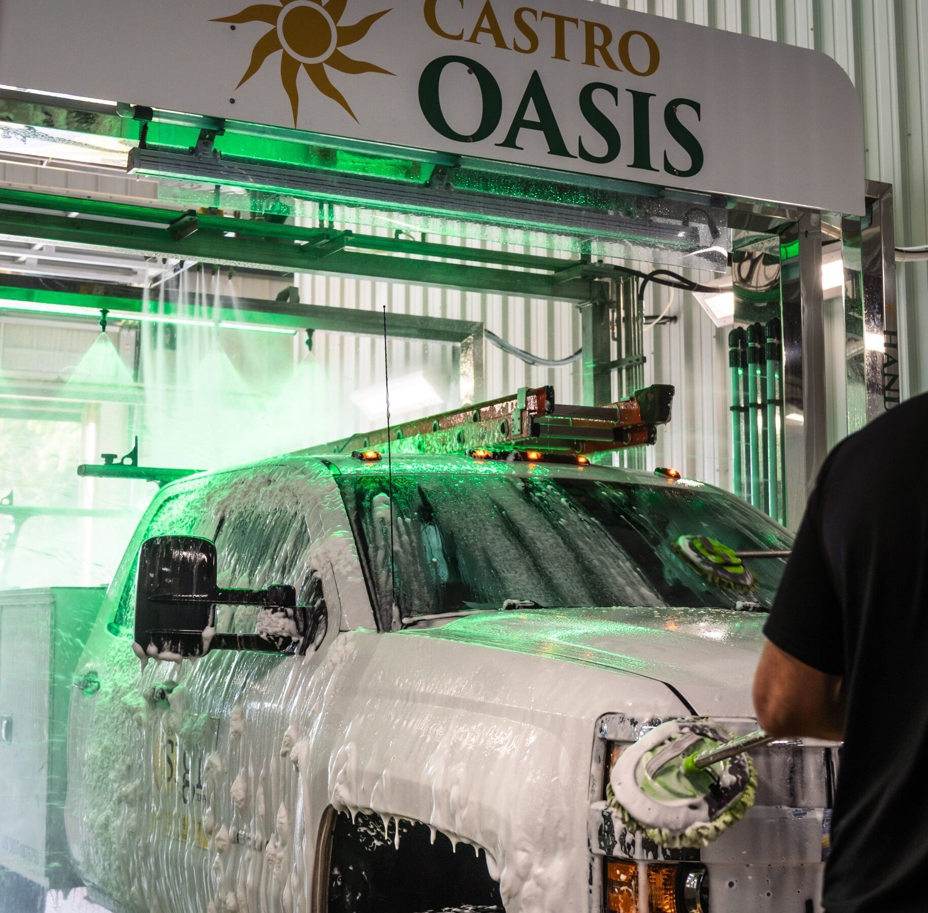 A person hand-washing a white truck covered in foam inside a Castro Oasis car wash bay.
