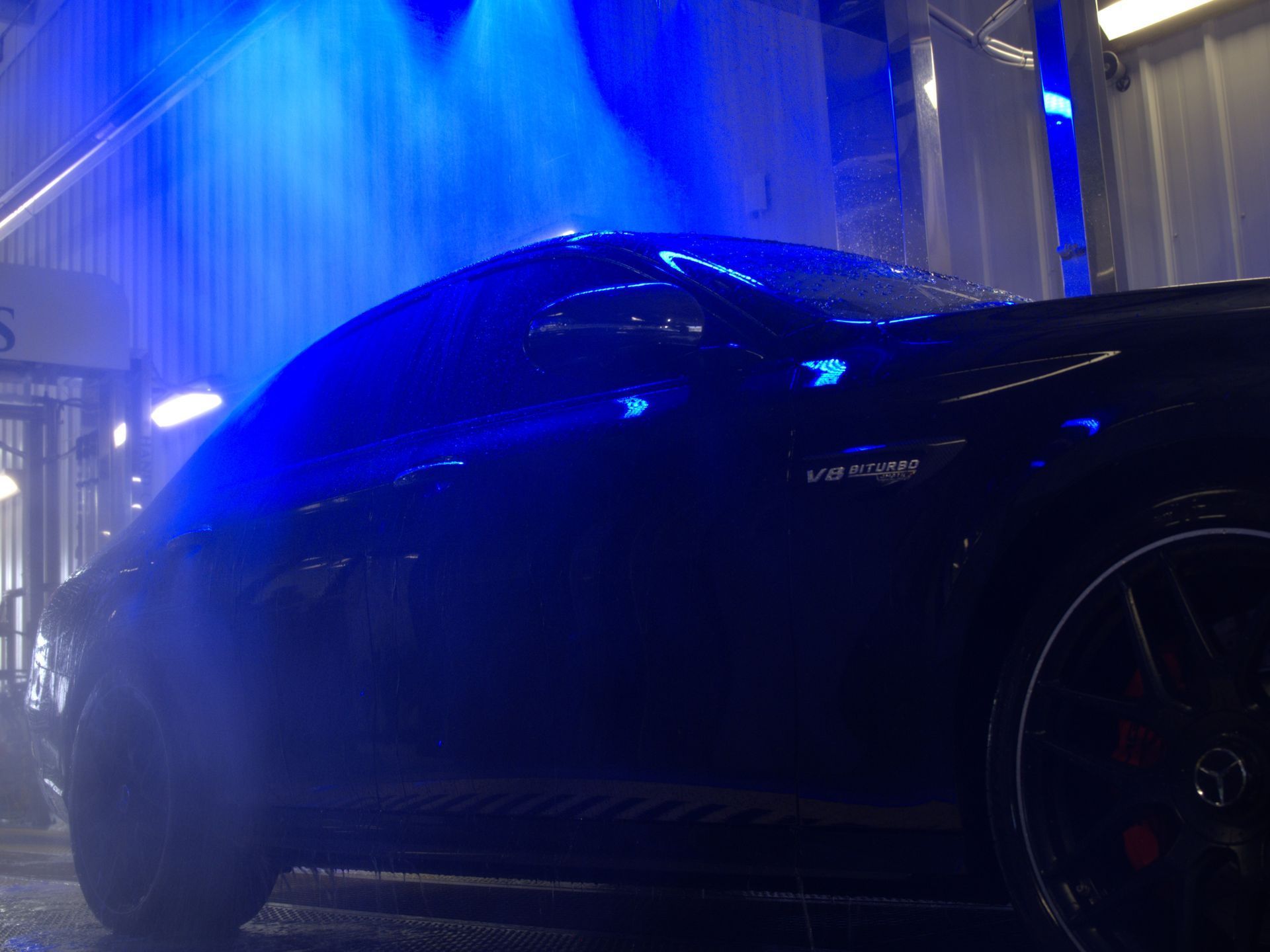 A dark car being cleaned in an automated car wash, illuminated by bright blue overhead lights.